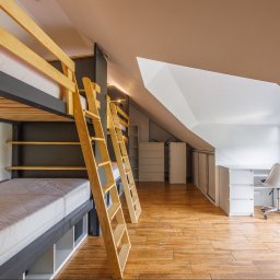 Opaliński Nieruchomości - Pokój na poddaszu z drewnianą podłogą, łóżkiem piętrowym z drabinkami i białym biurkiem z krzesłem, oświetlony punktowymi lampami.