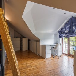 Opaliński Nieruchomości - Pokój na poddaszu z łóżkiem piętrowym, biurkiem przy oknie i zabudową szaf wnękowych, drewniana podłoga, niebieskie zasłony.