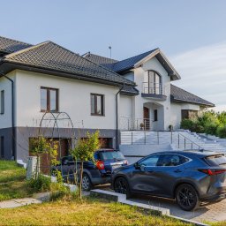 Opaliński Nieruchomości - Dwupiętrowy dom z szarą dachówką, białymi ścianami i dwoma zaparkowanymi samochodami na podjeździe z kostki brukowej.