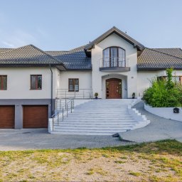 Opaliński Nieruchomości - Dwupiętrowy dom z garażem i białymi schodami prowadzącymi do wejścia, widoczny samochód osobowy zaparkowany obok