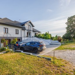 Opaliński Nieruchomości - Dwukondygnacyjny dom z szarym dachem, białymi ścianami i schodami wejściowymi, z dwoma samochodami zaparkowanymi na podjeździe i bujną zielenią w tle.