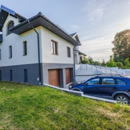 Opaliński Nieruchomości - Dwupiętrowy dom z szarym dachem i dwoma samochodami zaparkowanymi na podjeździe, widok z dołu na trawnik