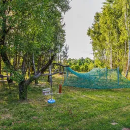 Zielony ogród z huśtawką na drzewie i siatką do leżenia, otoczony drzewami liściastymi i ścieżką z kamieni.