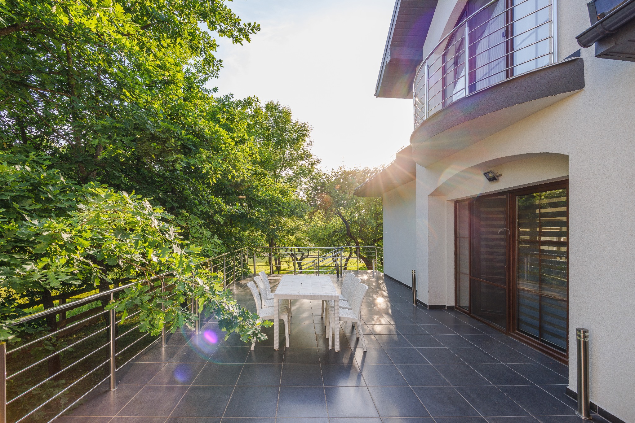 Przestronny taras z szarymi płytkami, białym stołem i krzesłami, otoczony zielenią drzew, z widokiem na ogród, w nowoczesnym domu z zaokrąglonym balkonem.