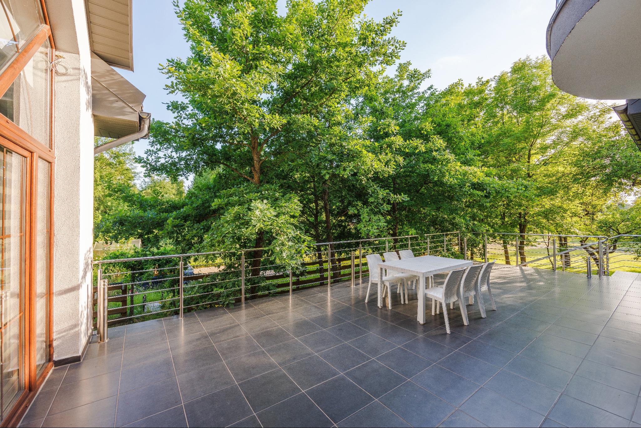 Przestronny taras z szarymi płytkami, białym stołem i krzesłami, otoczony zielenią drzew i metalową balustradą.