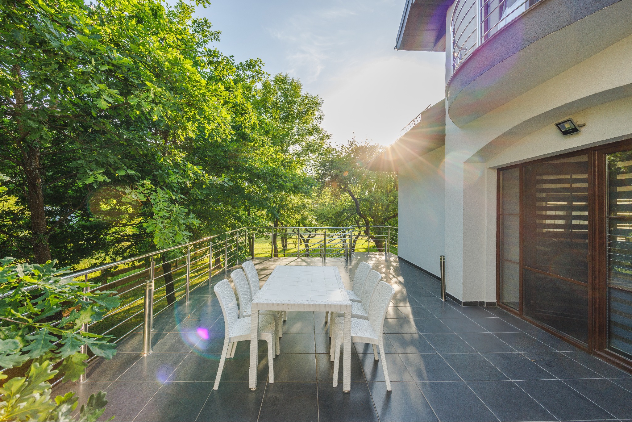 Biały stół i krzesła rattanowe na przestronnym tarasie z widokiem na zieleń, promienie słońca przebijają przez drzewa.