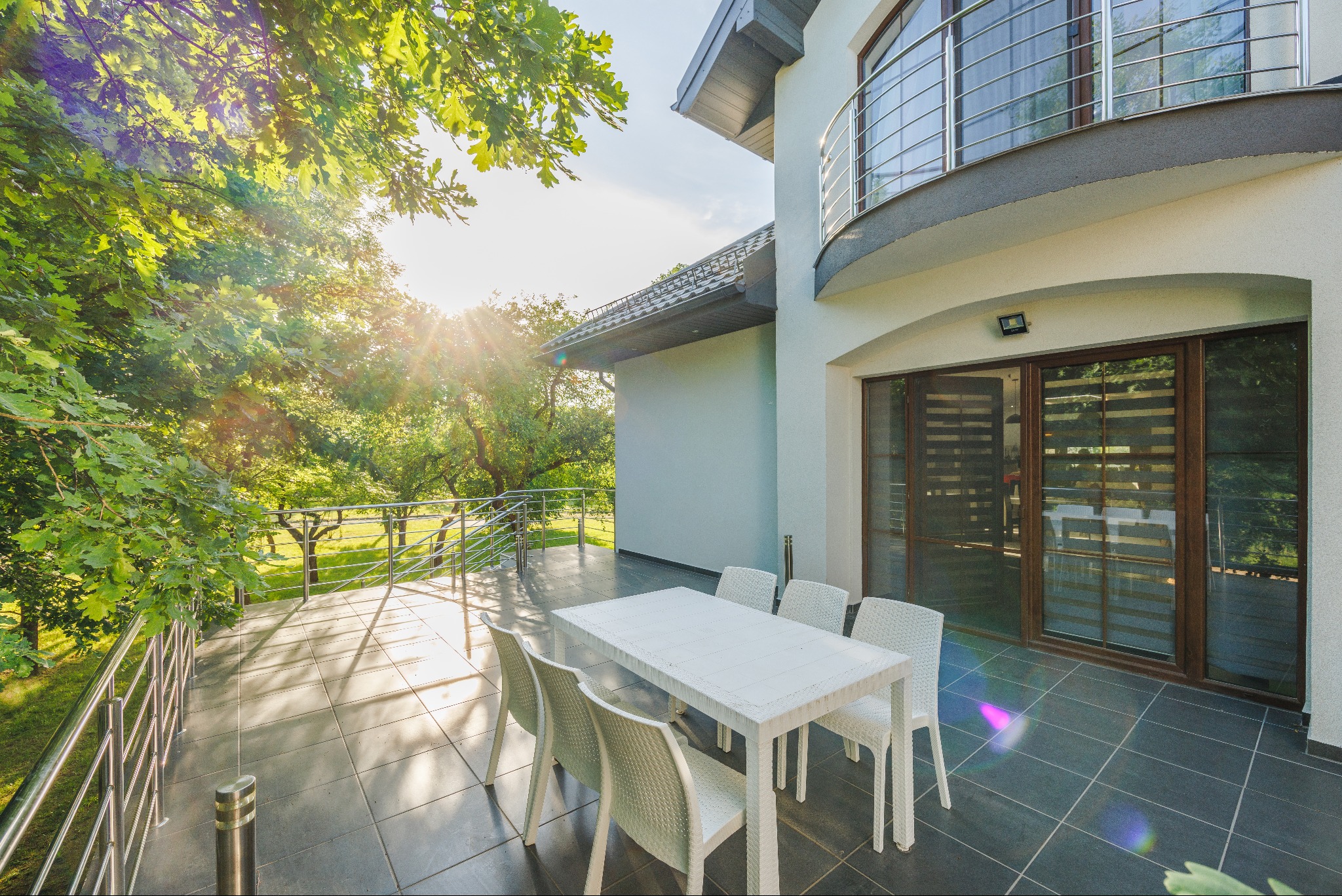 Nowoczesny taras z białym stołem i krzesłami z tworzywa, z widokiem na zielony ogród w słoneczny dzień.