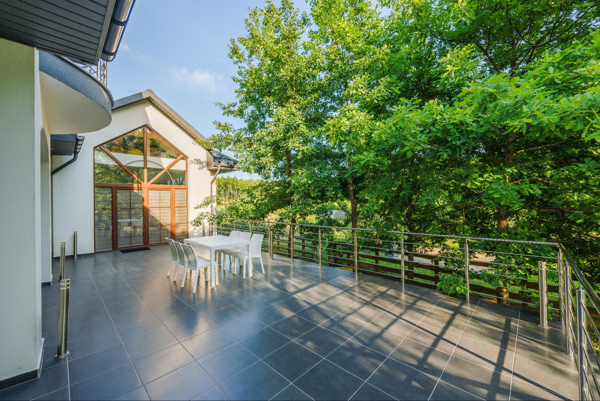 Przestronny taras z ciemnymi płytkami, białym stołem i krzesłami, widok na zieleń z metalową balustradą, nowoczesny budynek z dużym oknem trójkątnym.