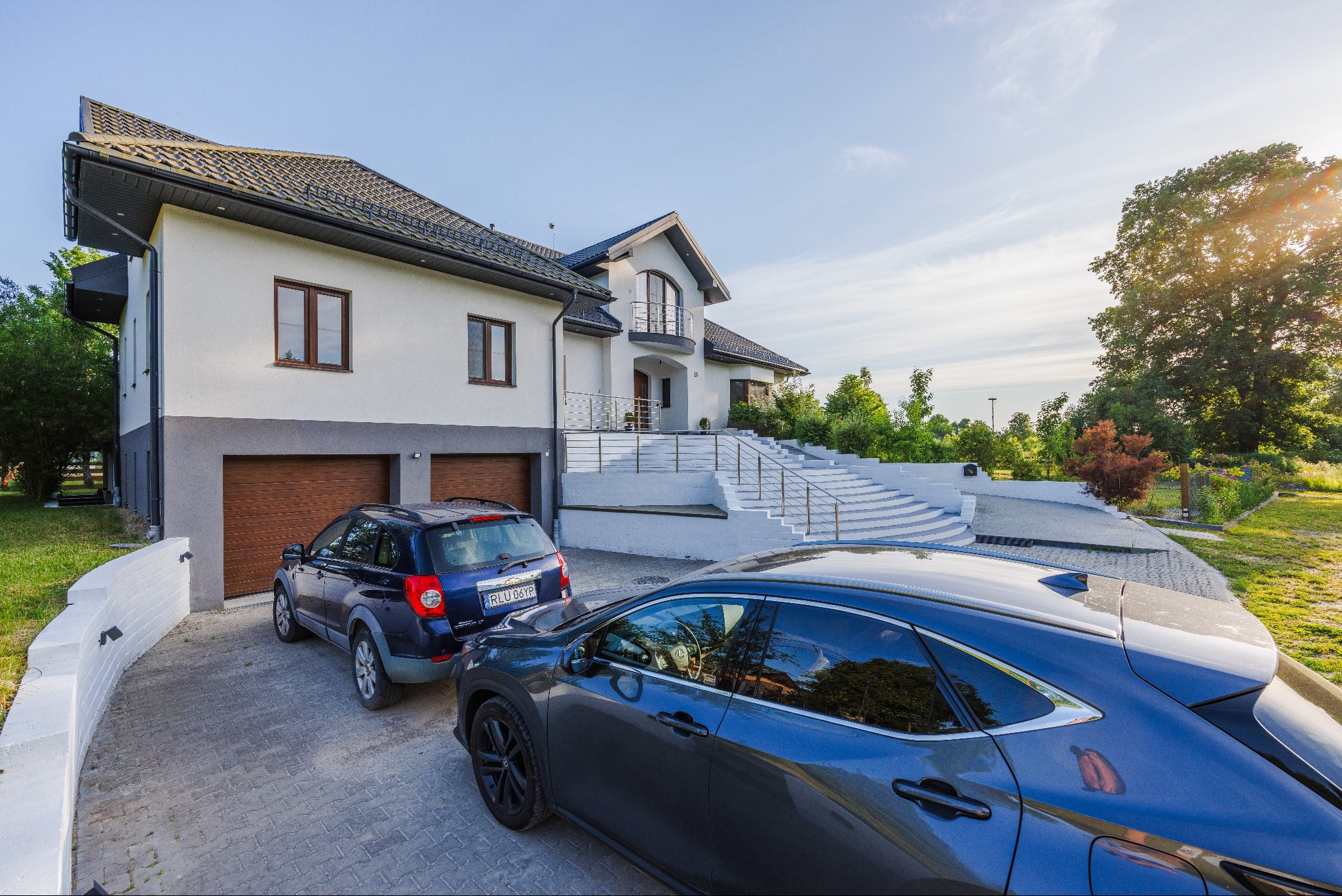 Dwupiętrowy dom z białymi ścianami i szarym cokołem, dwoma garażami z drewnianymi bramami, schodami prowadzącymi do wejścia oraz dwoma zaparkowanymi samochodami na podjeździe z kostki brukowej.