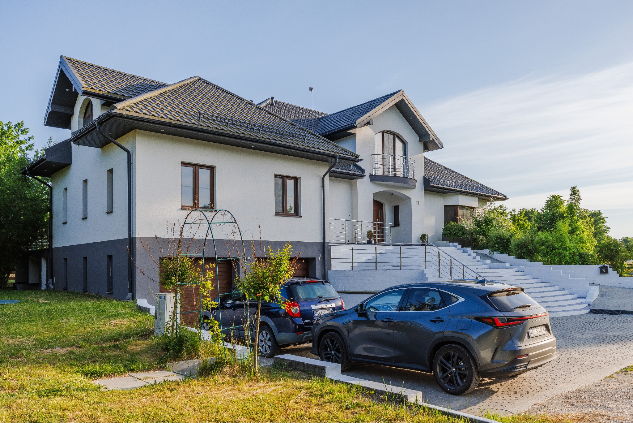 Dwupiętrowy dom z szarą dachówką, białymi ścianami i dwoma zaparkowanymi samochodami na podjeździe z kostki brukowej.