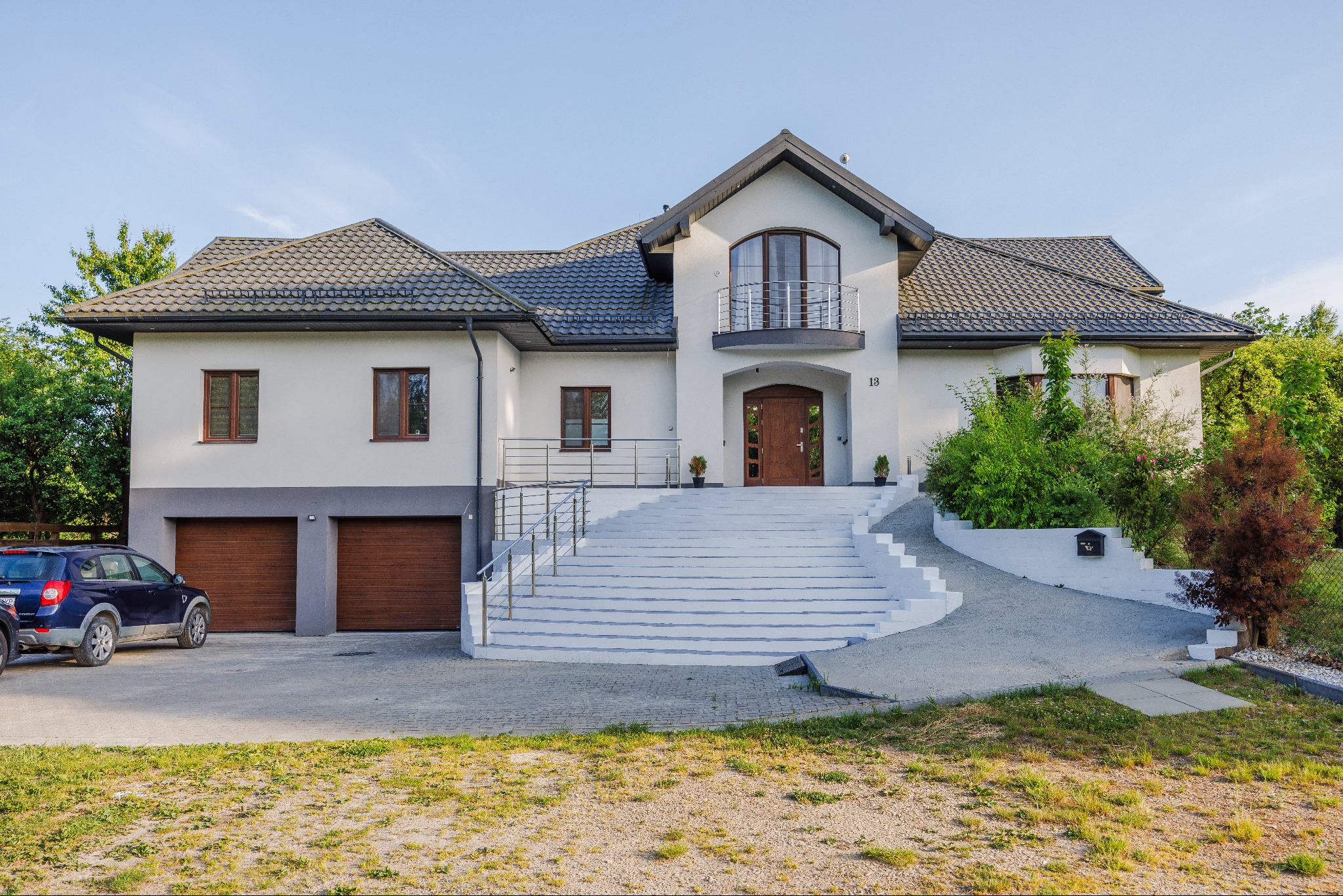 Dwupiętrowy dom z garażem i białymi schodami prowadzącymi do wejścia, widoczny samochód osobowy zaparkowany obok