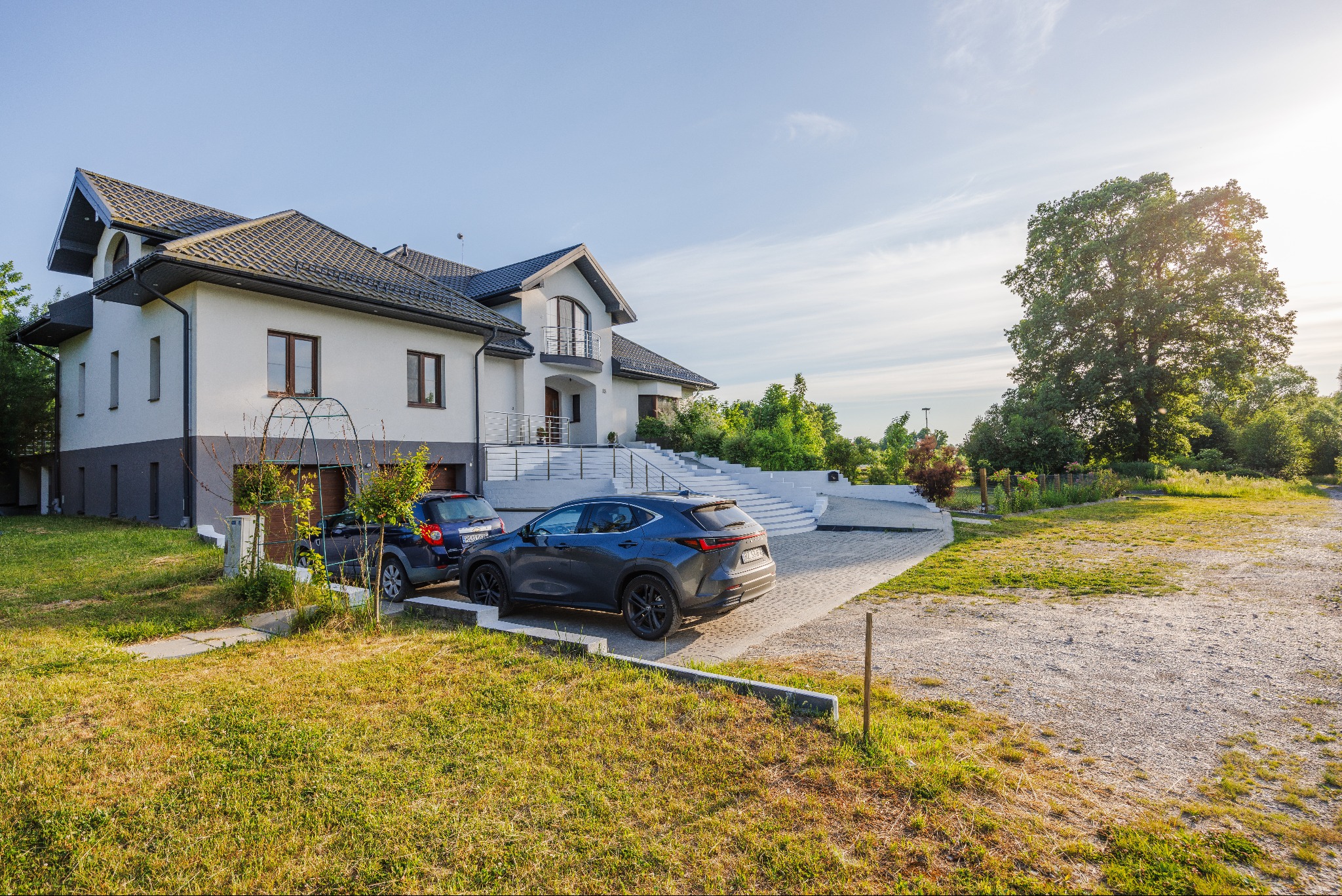 Dwukondygnacyjny dom z szarym dachem, białymi ścianami i schodami wejściowymi, z dwoma samochodami zaparkowanymi na podjeździe i bujną zielenią w tle.