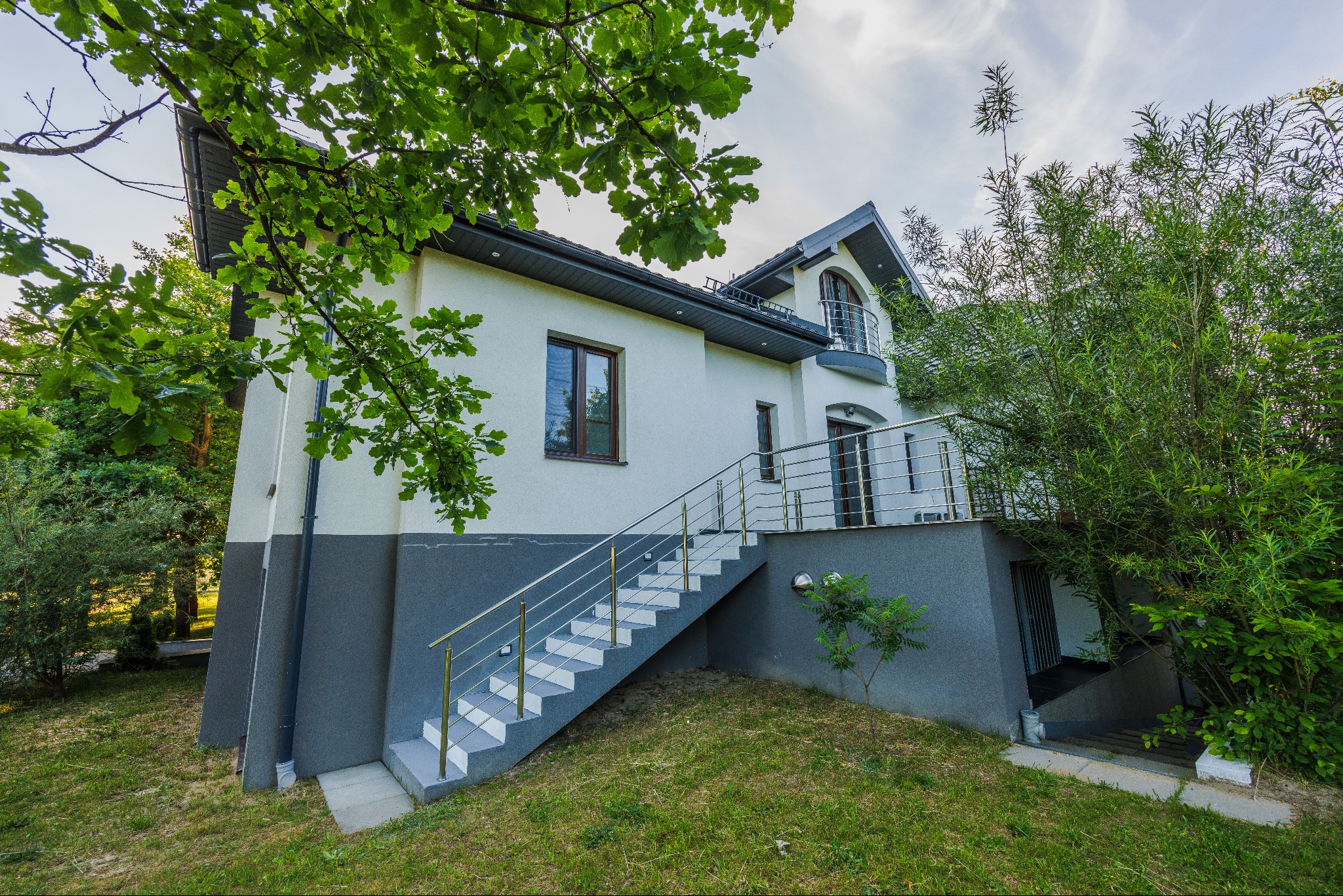 Dwukondygnacyjny dom z zewnętrznymi schodami prowadzącymi na balkon, z jasną elewacją i szarą podmurówką, otoczony zielenią i drzewami.