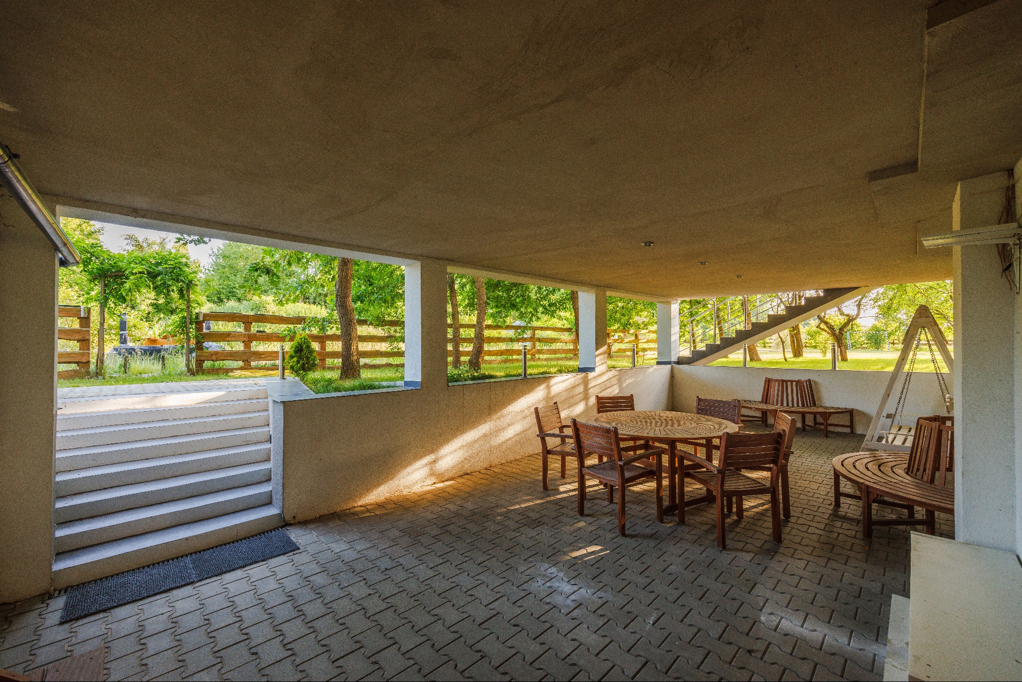 Zadaszony taras z drewnianymi meblami ogrodowymi, widok na zielony ogród z drewnianym płotem i schodami prowadzącymi na zewnątrz.