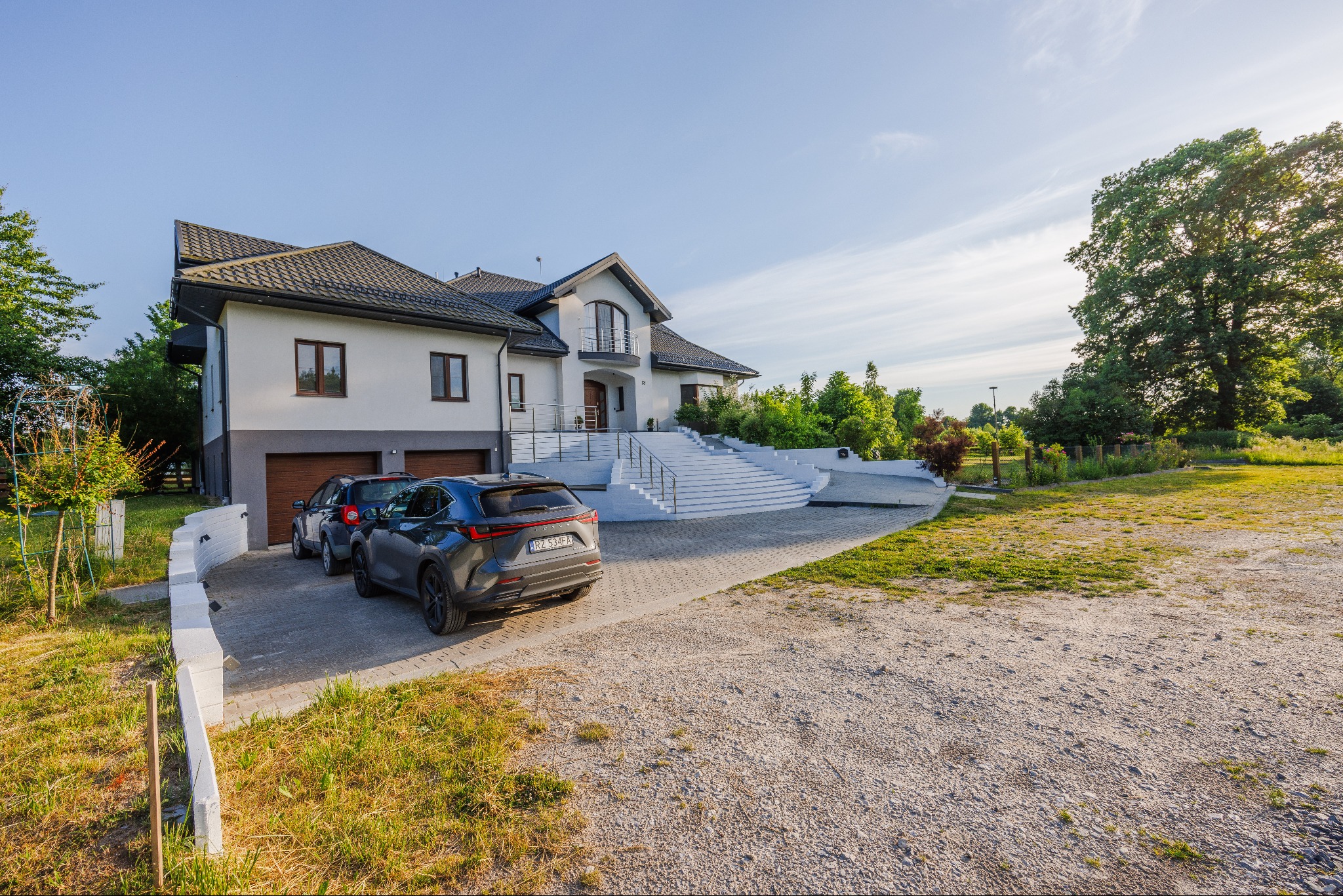 Dwukondygnacyjny dom z garażem i białymi schodami prowadzącymi do wejścia, z zaparkowanymi dwoma samochodami na podjeździe.