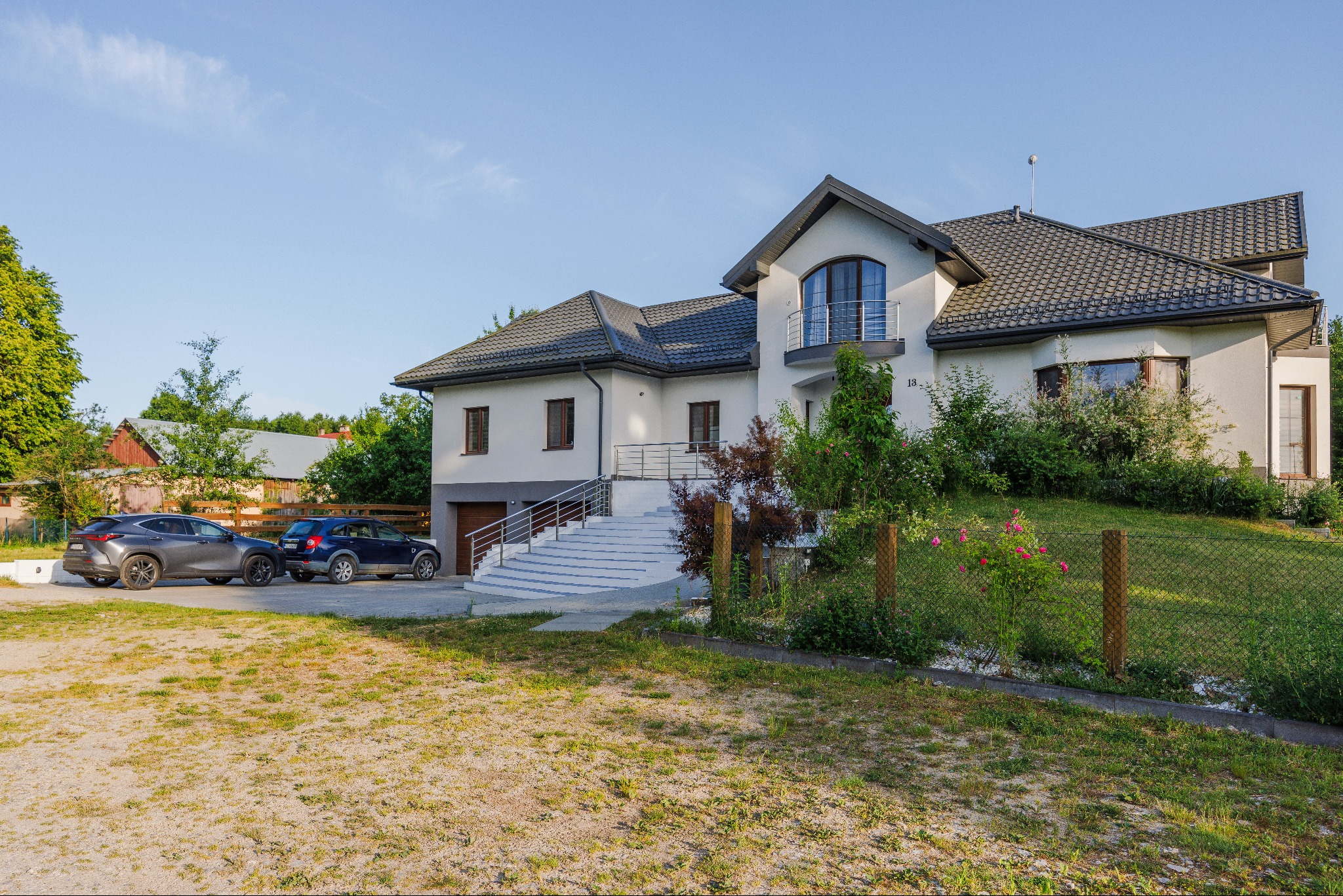 Nowoczesny dom jednorodzinny z szarą dachówką i białymi ścianami, z balkonem z metalową balustradą, dwoma samochodami zaparkowanymi przed domem i ogrodem z ogrodzeniem z siatki.