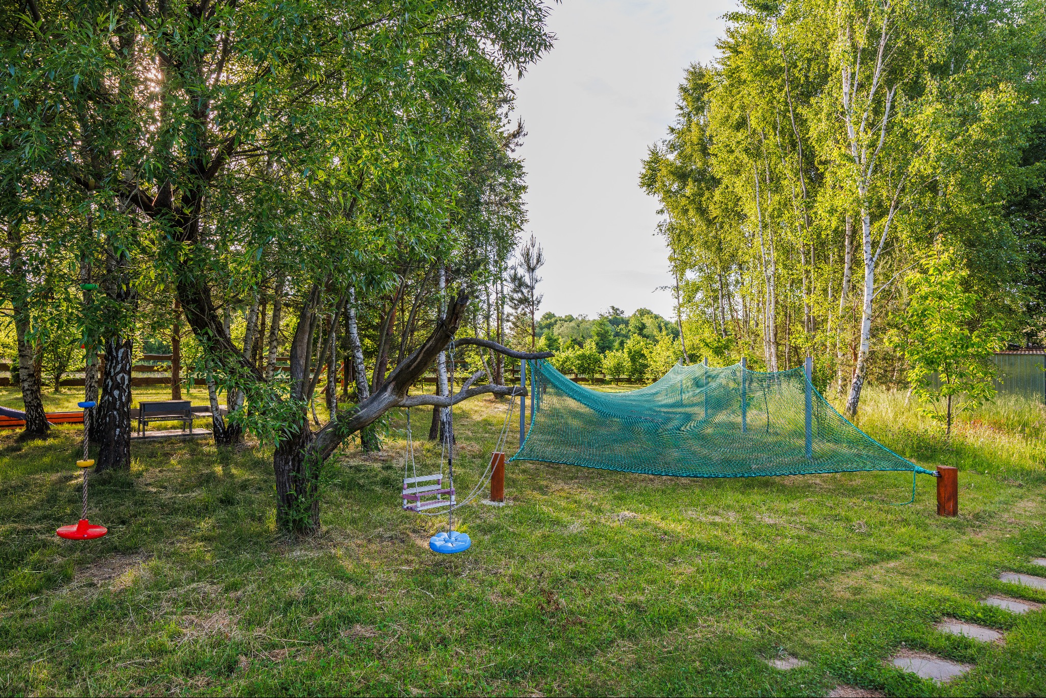 Zielony ogród z huśtawką na drzewie i siatką do leżenia, otoczony drzewami liściastymi i ścieżką z kamieni.