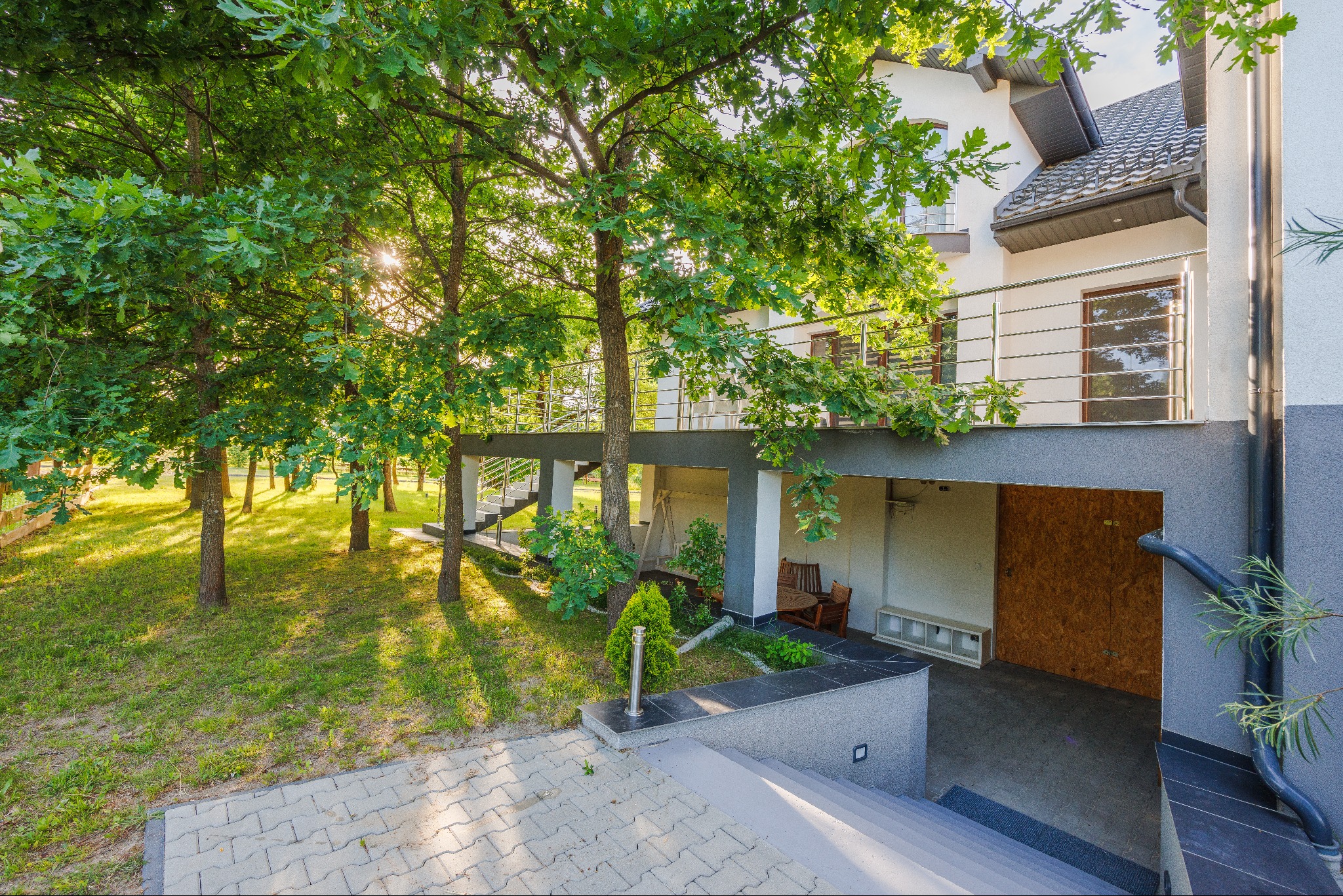 Dwupiętrowy dom z balkonem i schodami zewnętrznymi, otoczony zielenią, z widoczną częścią garażu z zamkniętymi drzwiami z płyty OSB.