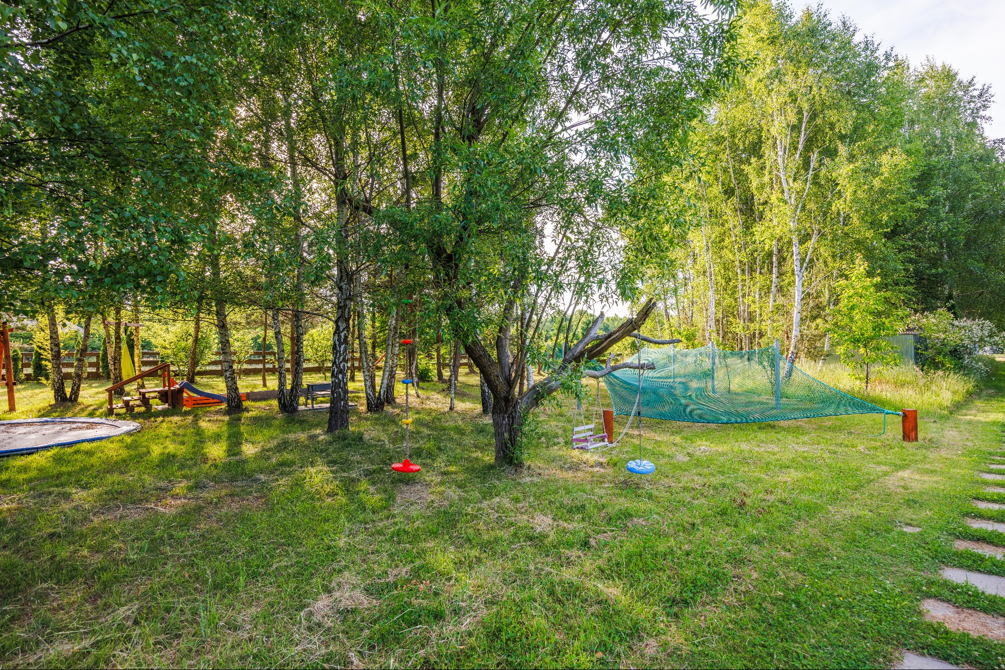 Zielony ogród z placem zabaw: trampolina, huśtawki na drzewach i siatka do wspinaczki, otoczone drzewami i ścieżką z kamieni.