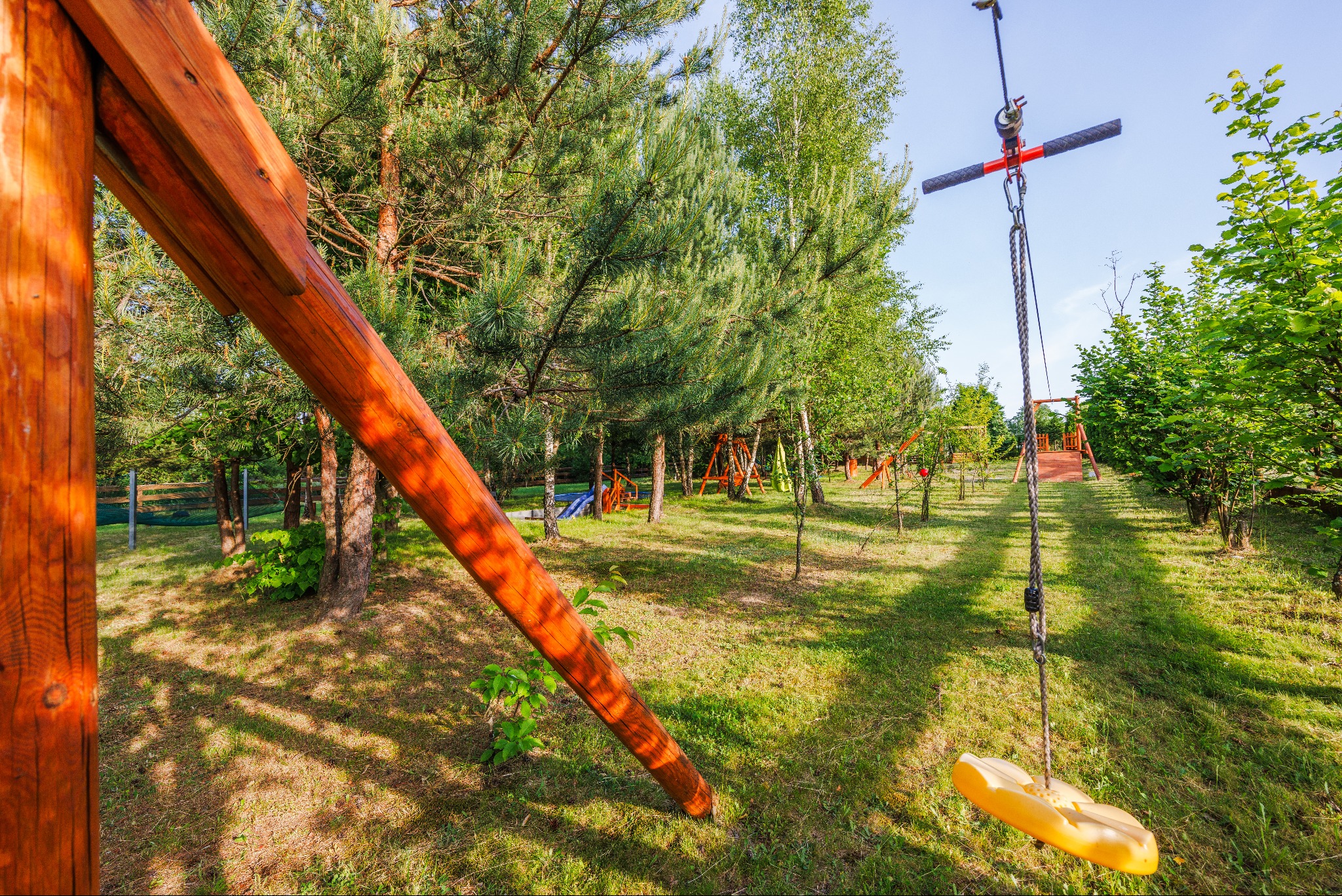 Drewniany plac zabaw z huśtawką linową i siedziskiem w kształcie motyla, otoczony zielenią drzew i trawnikiem, w oddali widoczne inne elementy placu zabaw.