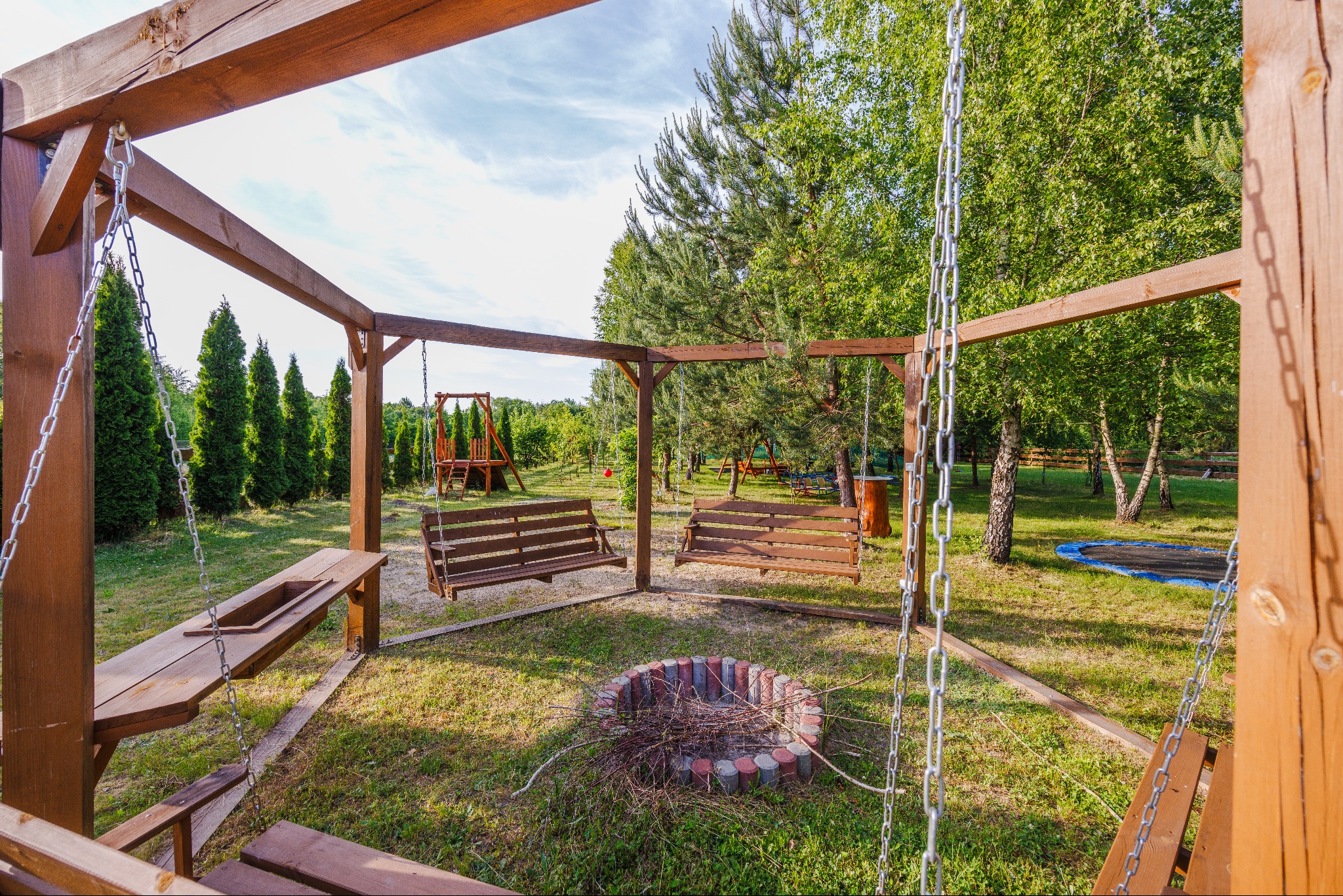 Drewniana konstrukcja z huśtawkami łańcuchowymi i miejscem na ognisko z cegieł w Rzeszowie, otoczona zielenią drzew i krzewów, widoczny również stół z wbudowanym miejscem na grill.