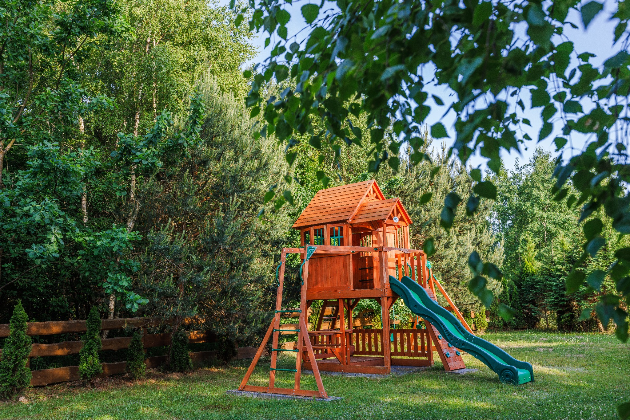 Drewniany plac zabaw z domkiem, zjeżdżalnią i drabinką w ogrodzie, otoczony zielenią drzew i krzewów, widok z perspektywy lekko z dołu poprzez gałęzie drzewa.