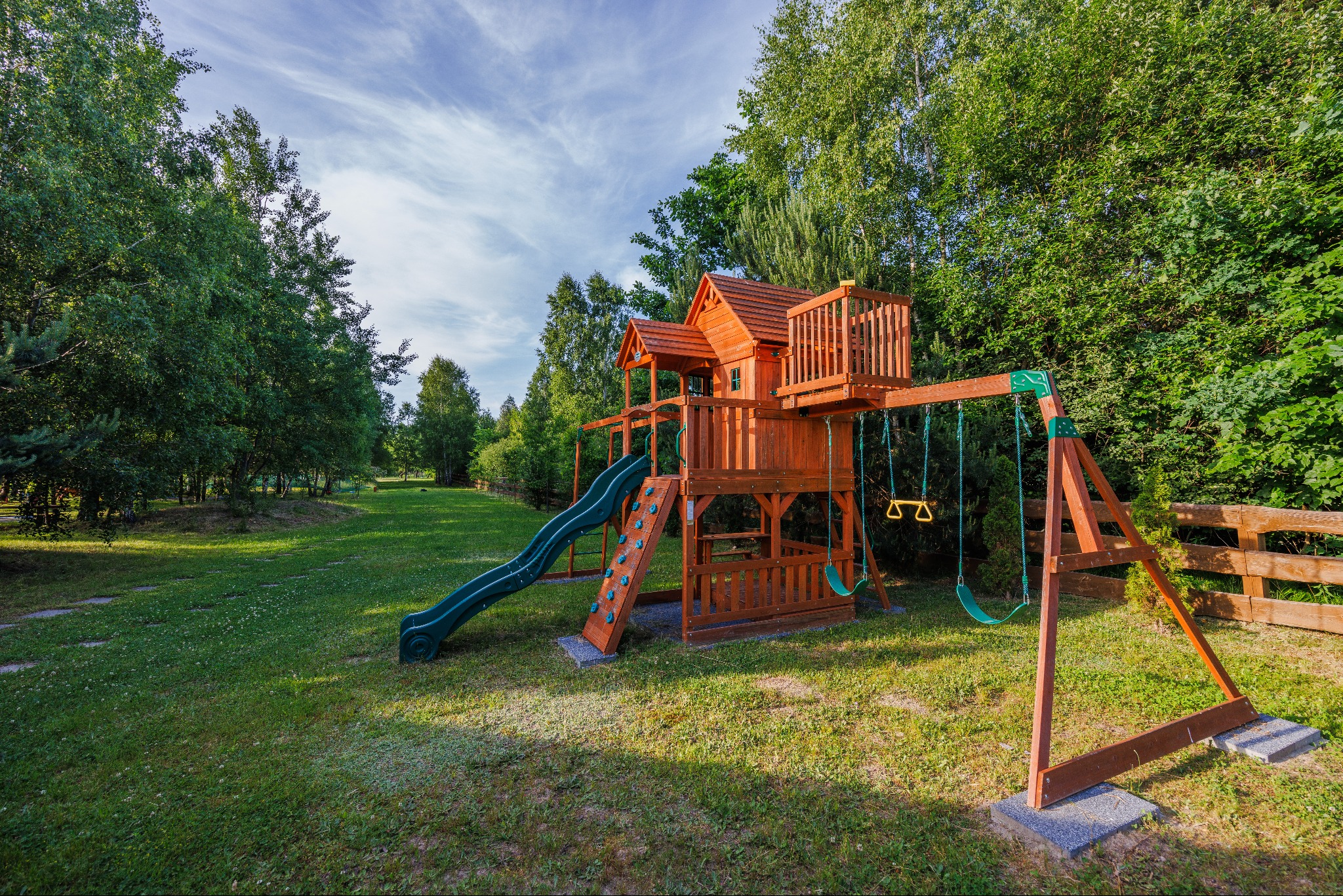 Drewniany plac zabaw z domkiem, zjeżdżalnią i huśtawkami na trawniku w otoczeniu drzew.