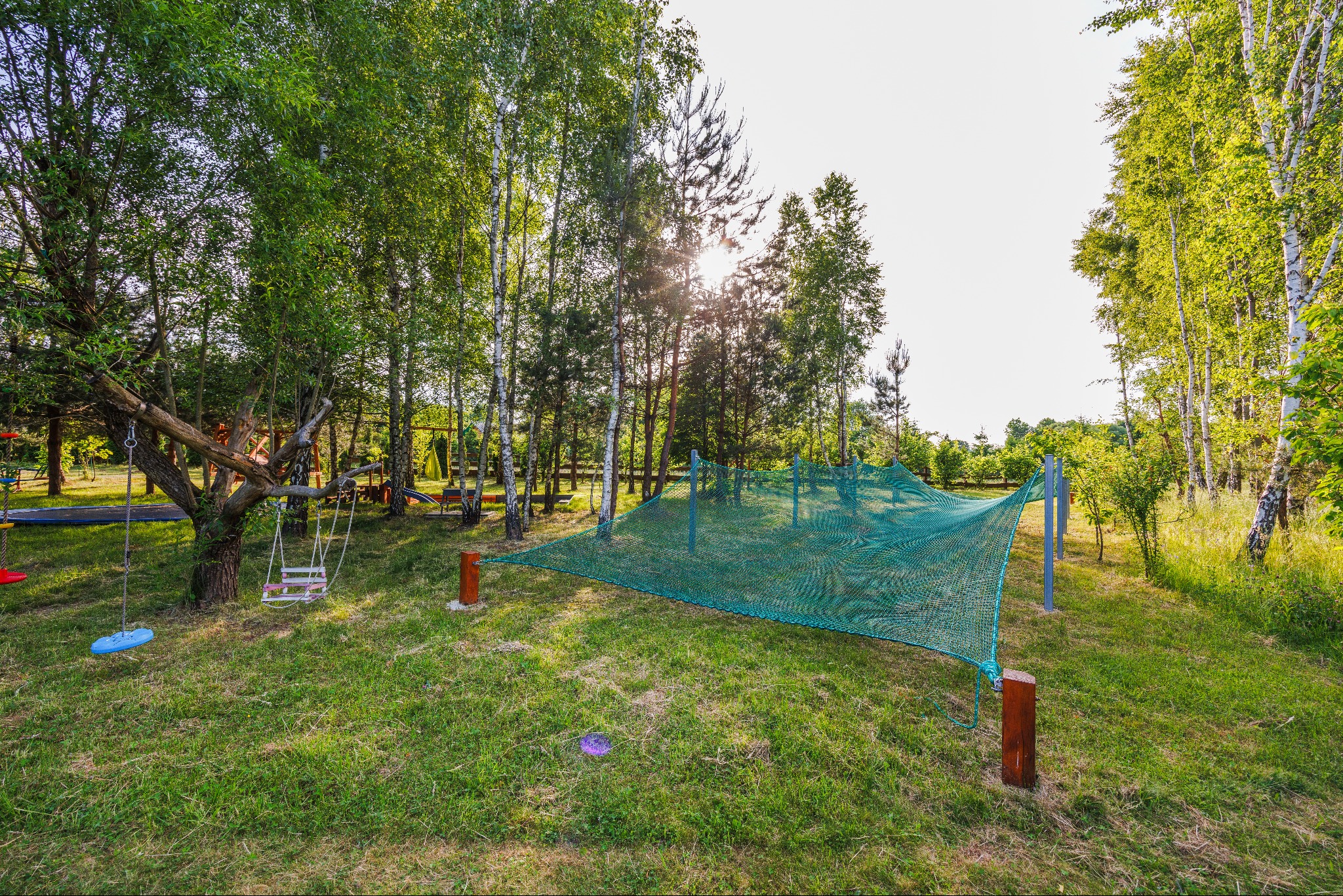 Zielony plac zabaw z siatką zabezpieczającą, huśtawkami i drzewami w tle, oświetlony przez słońce.