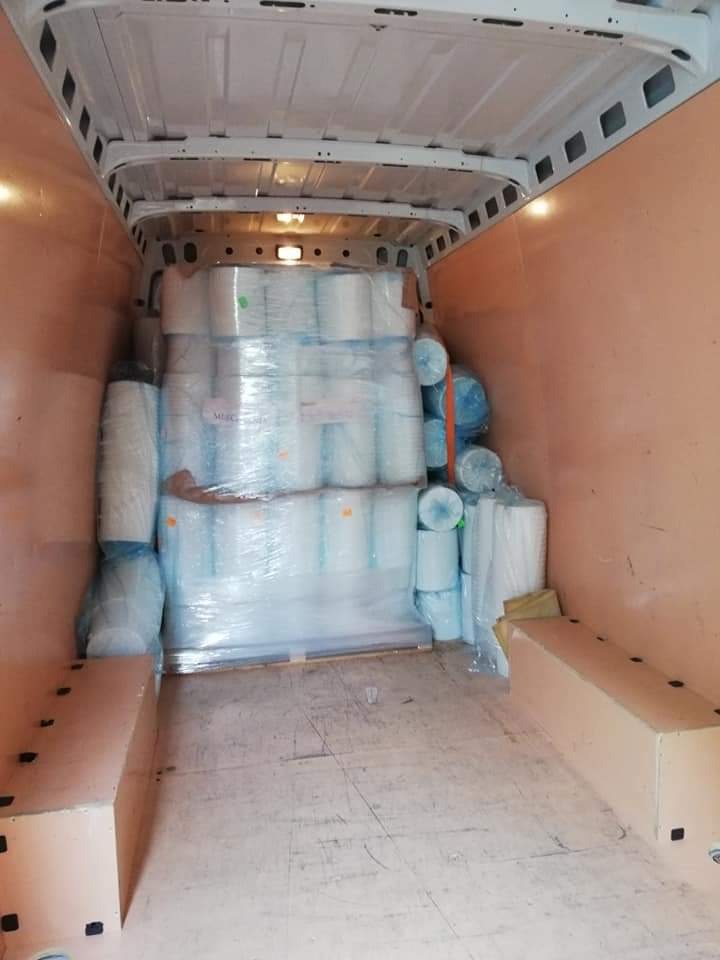 Wnętrze furgonetki załadowane rolkami białego materiału, zabezpieczonymi folią stretch, widoczne drewniane skrzynie po bokach ładunku.