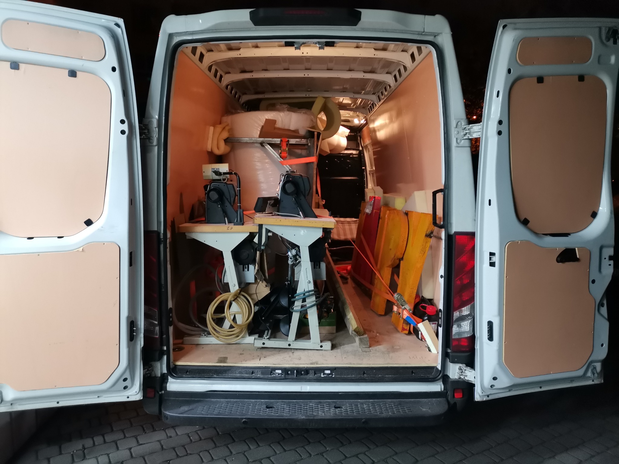 Wnętrze otwartego furgonetki wypełnionej materiałami tapicerskimi, w tym belami pianki i dwoma maszynami do szycia na stołach, przygotowane do transportu.