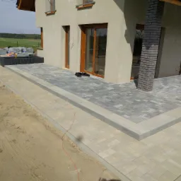 Nowoczesny taras z szarej i beżowej kostki brukowej, wykończony obrzeżami, z widocznymi elementami domu w tle i stertą kostki brukowej na trawniku.