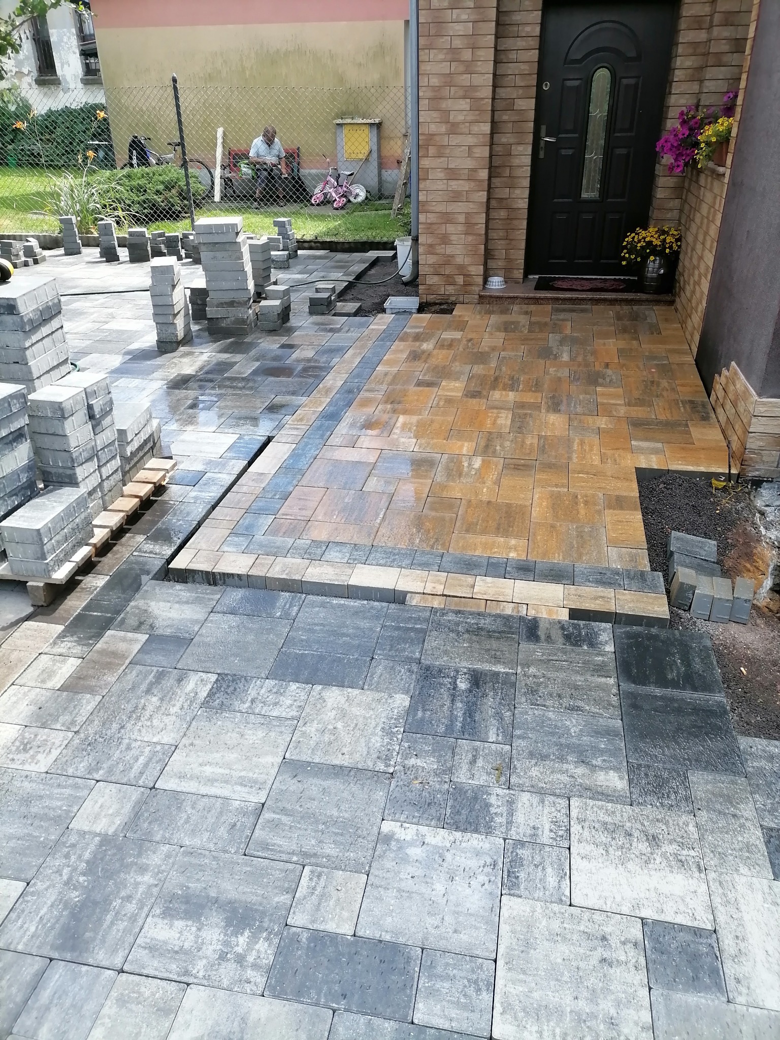 W trakcie układania wielokolorowej kostki brukowej przed domem, z widocznymi stertami kostki i fragmentami już ułożonej nawierzchni.