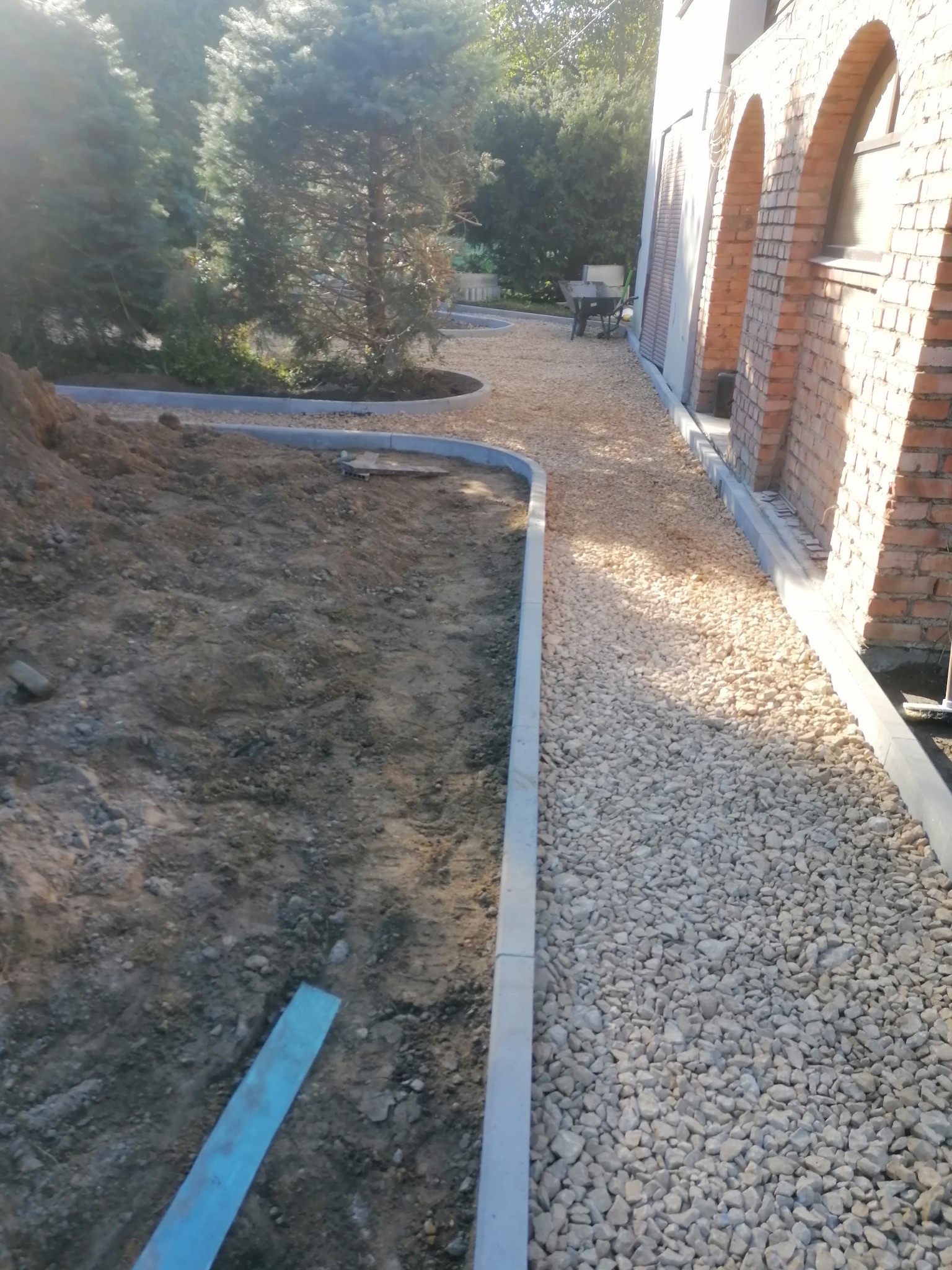 Przygotowanie podłoża: wyrównany teren z częściowo ułożonym obrzeżem z szarego betonu i wysypanym białym grysem, widoczna sterta ziemi i elementy narzędzi budowlanych.