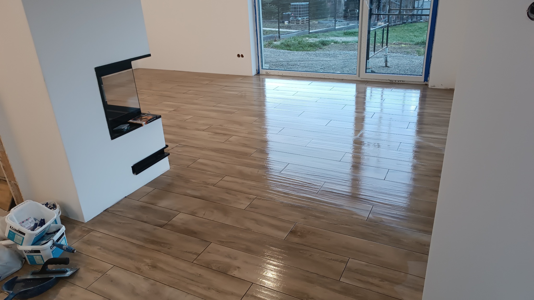Połyskująca podłoga z płytek imitujących drewno, ułożona w salonie z nowoczesnym kominkiem; widoczne wiaderka z zaprawą i narzędzia glazurnicze.
