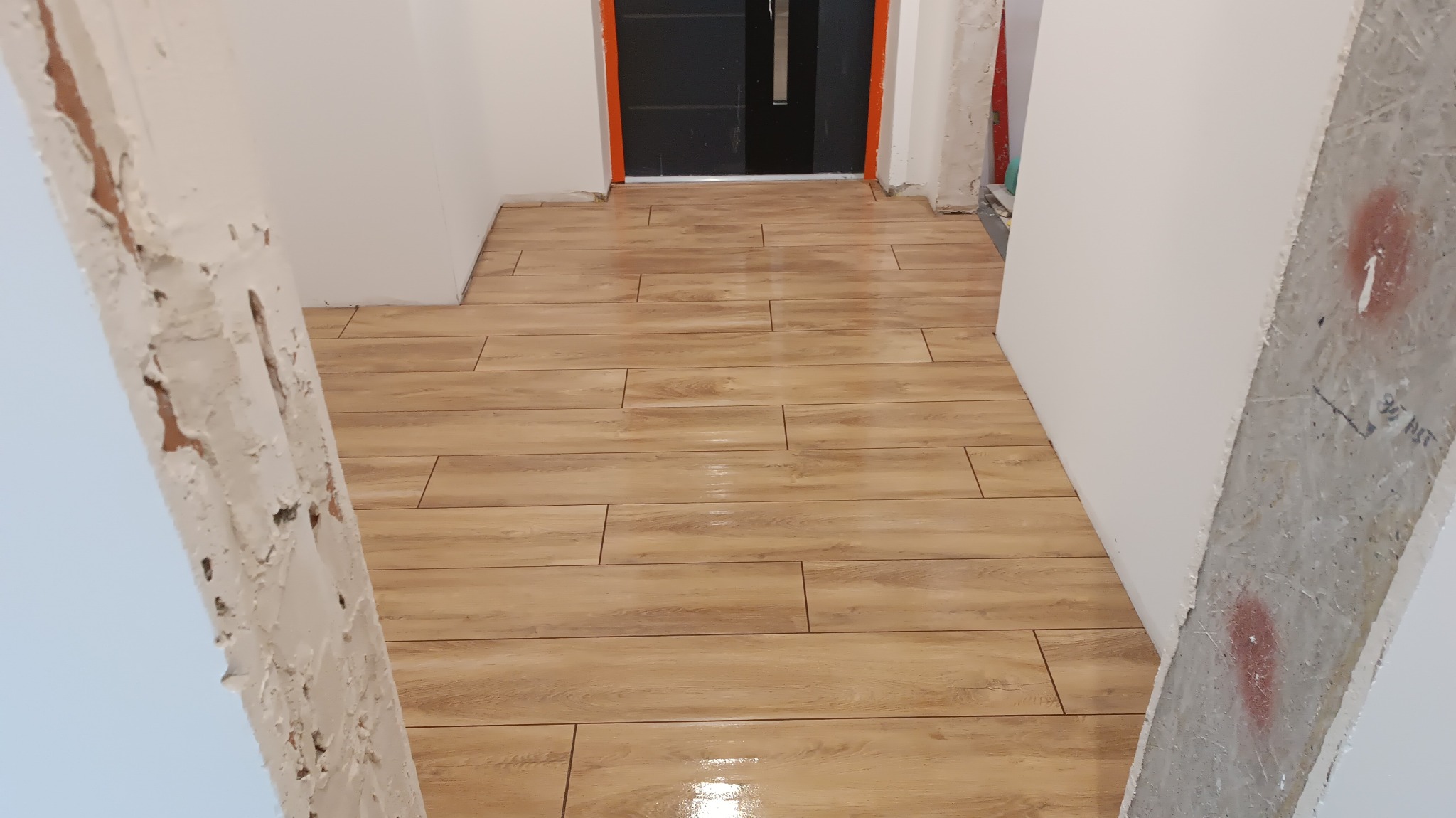 Ułożona podłoga z płytek imitujących drewno w korytarzu w trakcie remontu, widoczne surowe ściany i framuga drzwi z pomarańczowym akcentem.