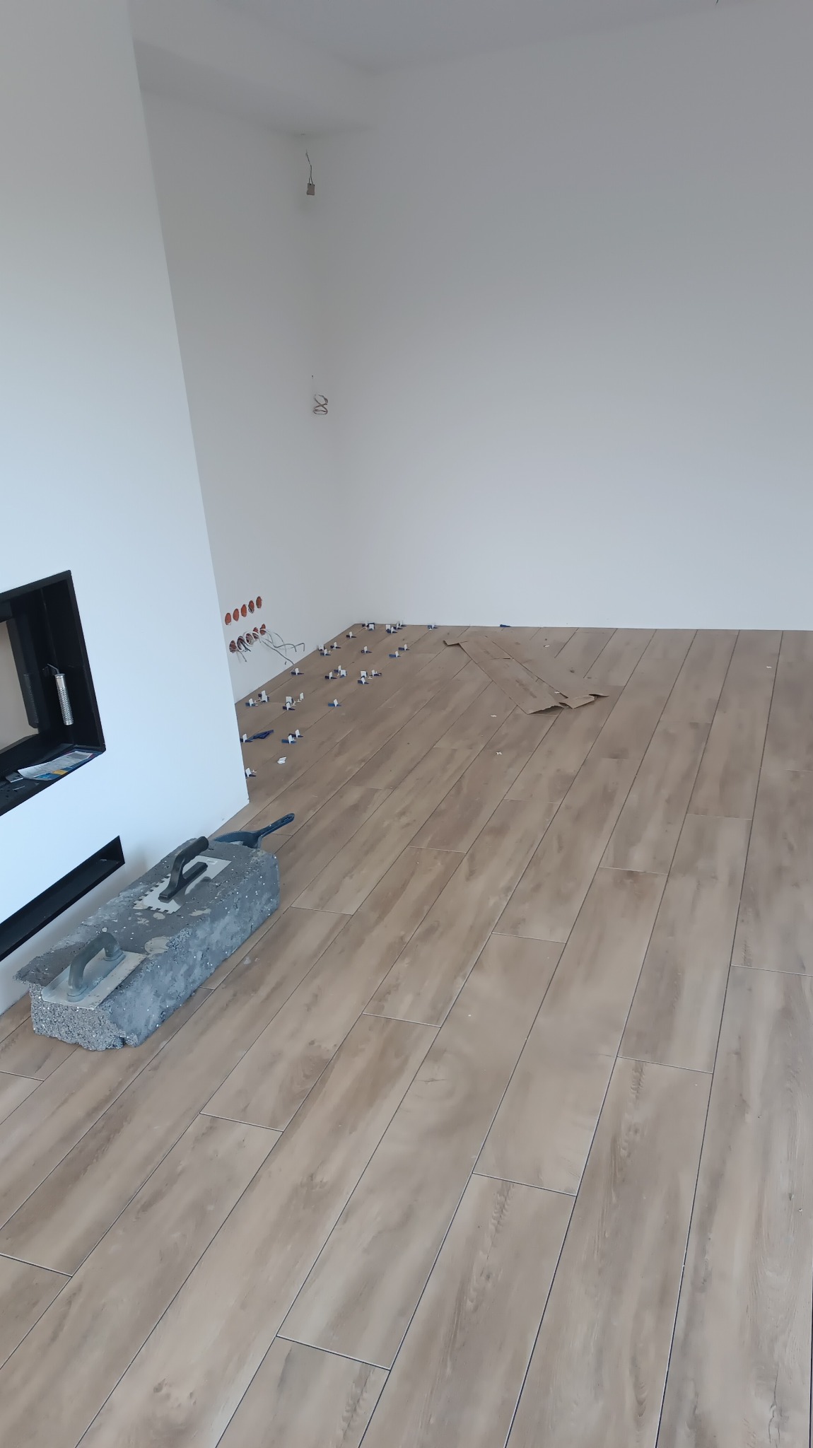 Układanie podłogi z płytek imitujących drewno w jasnym pomieszczeniu z kominkiem; widoczne krzyżyki dystansowe i narzędzia glazurnicze.