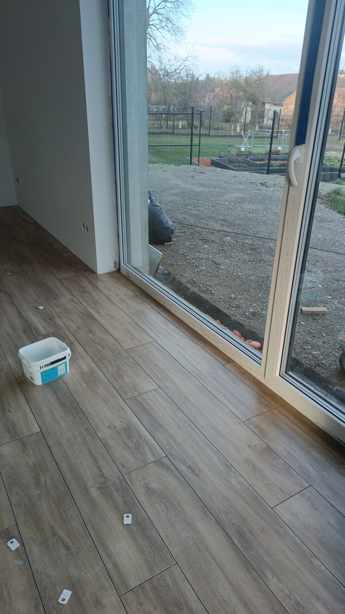 Ułożona podłoga z paneli imitujących drewno w nowym pomieszczeniu, widoczne białe separatory, wiadro z farbą i duże okno przesuwne z widokiem na ogród.