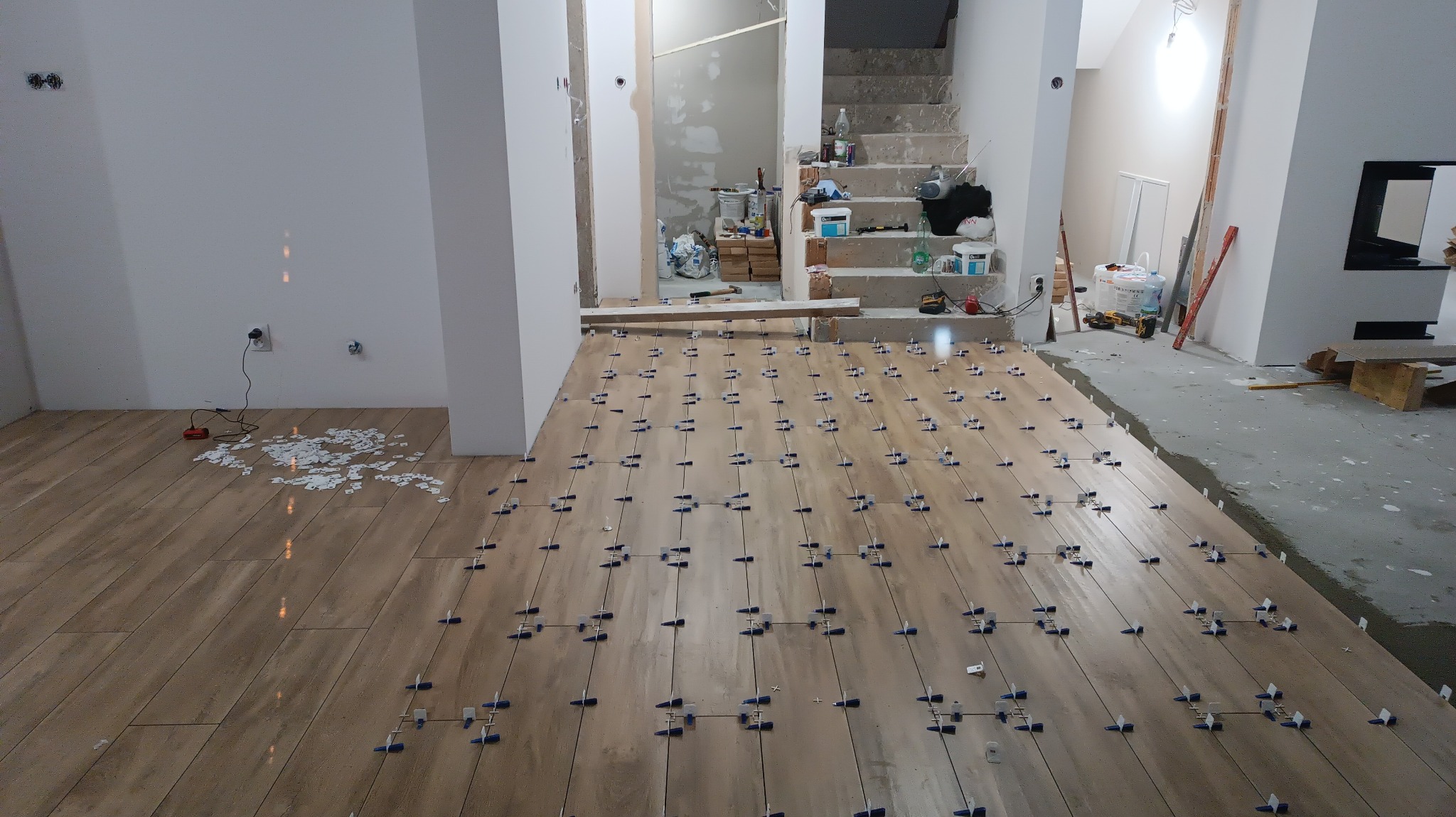 Układanie podłogi z płytek imitujących drewno, z użyciem niebieskich krzyżyków dystansowych, widok na schody i przygotowane materiały budowlane w tle.