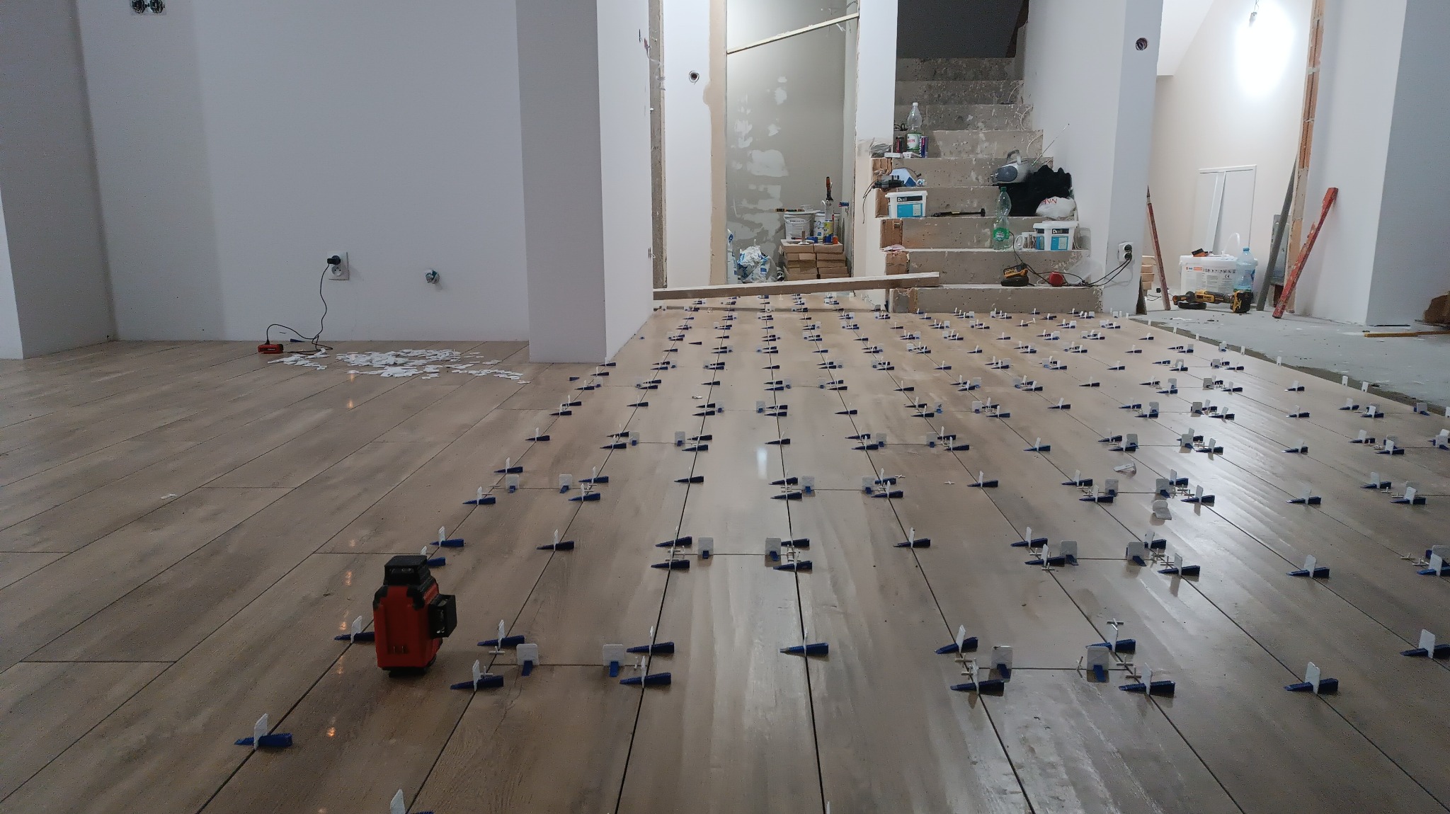 Proces układania płytek podłogowych z użyciem poziomicy laserowej i systemu poziomowania płytek w przestronnym wnętrzu, widoczne schody w tle.