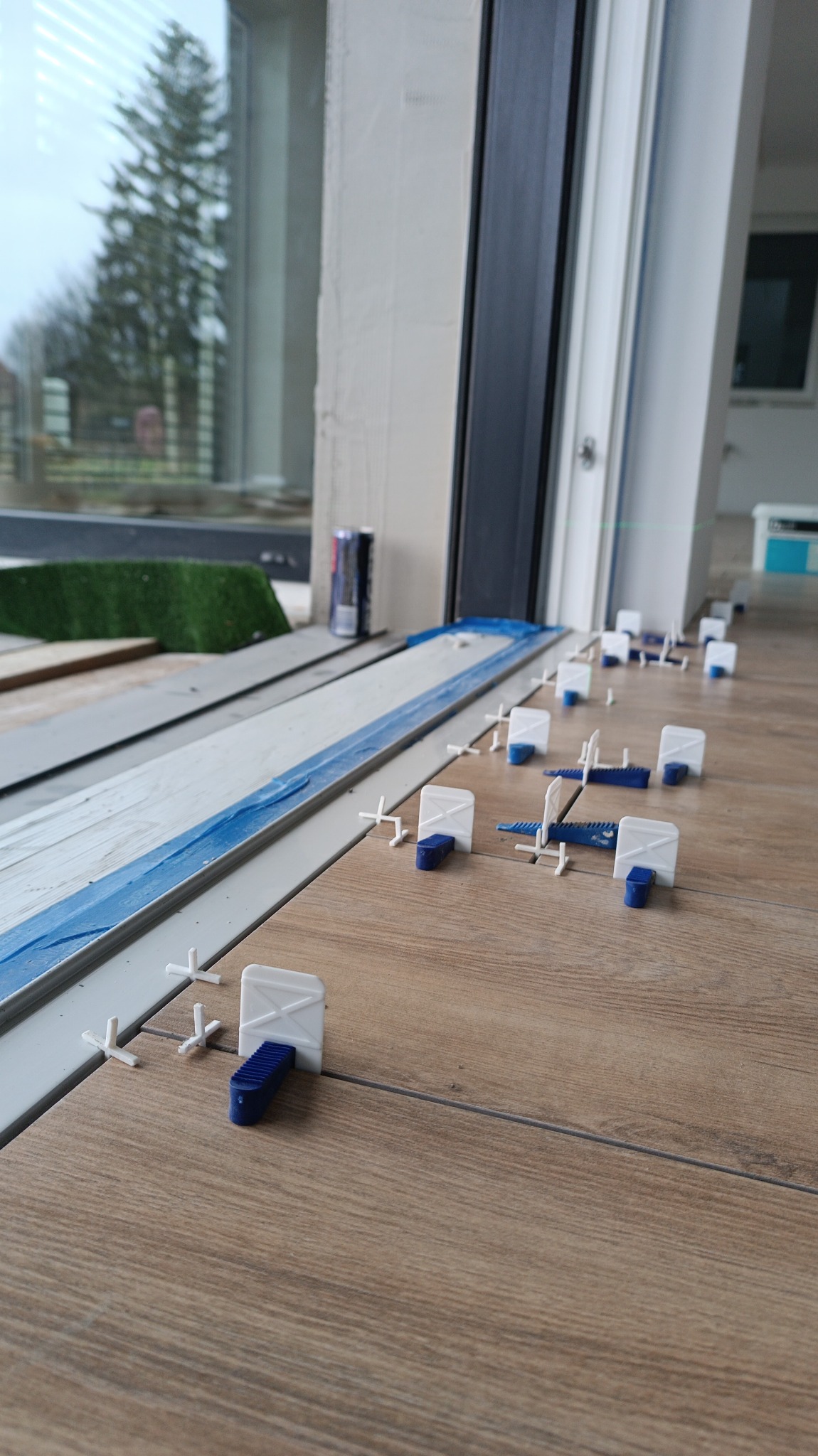 Ułożone płytki podłogowe imitujące drewno, z systemem poziomowania płytek (białe klipsy i niebieskie kliny) w trakcie montażu przy przesuwanych drzwiach balkonowych; widoczna puszka napoju w tle.