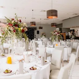 Uroczysta sala bankietowa z okrągłymi stołami nakrytymi białymi obrusami, szarymi krzesłami, kwiatowymi dekoracjami w wysokich wazonach, elegancką zastawą i nowoczesnymi lampami wiszącymi...