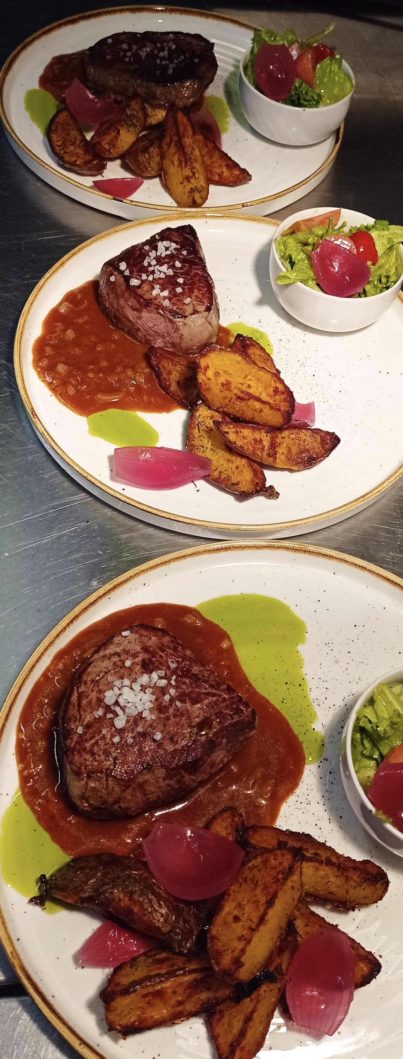 Dwa talerze z filetami wołowymi posypanymi solą, podanymi z pieczonymi ziemniakami, sosem i małą miseczką sałatki na metalowym blacie.