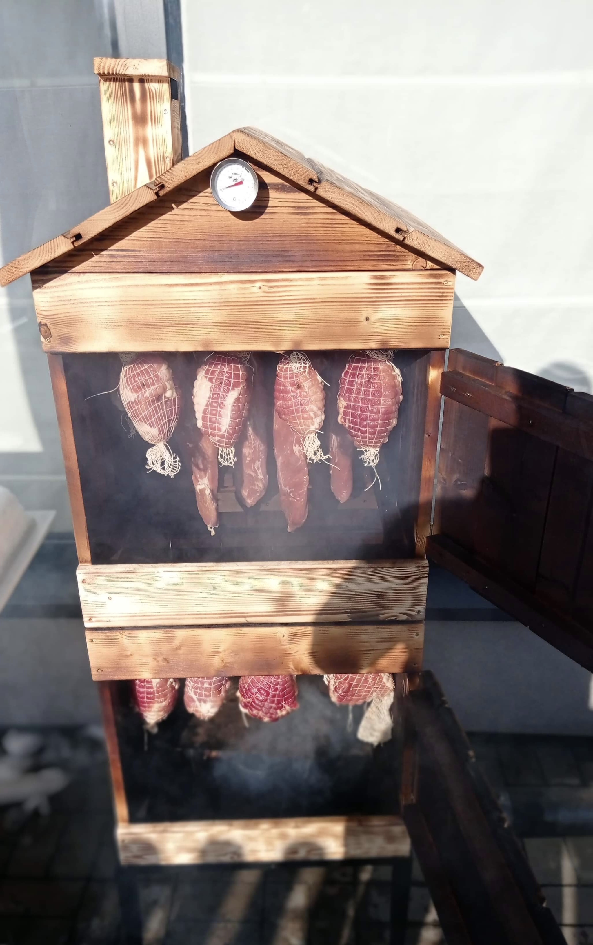 Wędzenie mięs w drewnianej wędzarni z termometrem, widoczne wędzone szynki i kiełbasy w siatkach, dym wewnątrz wędzarni, otwarte drzwiczki.