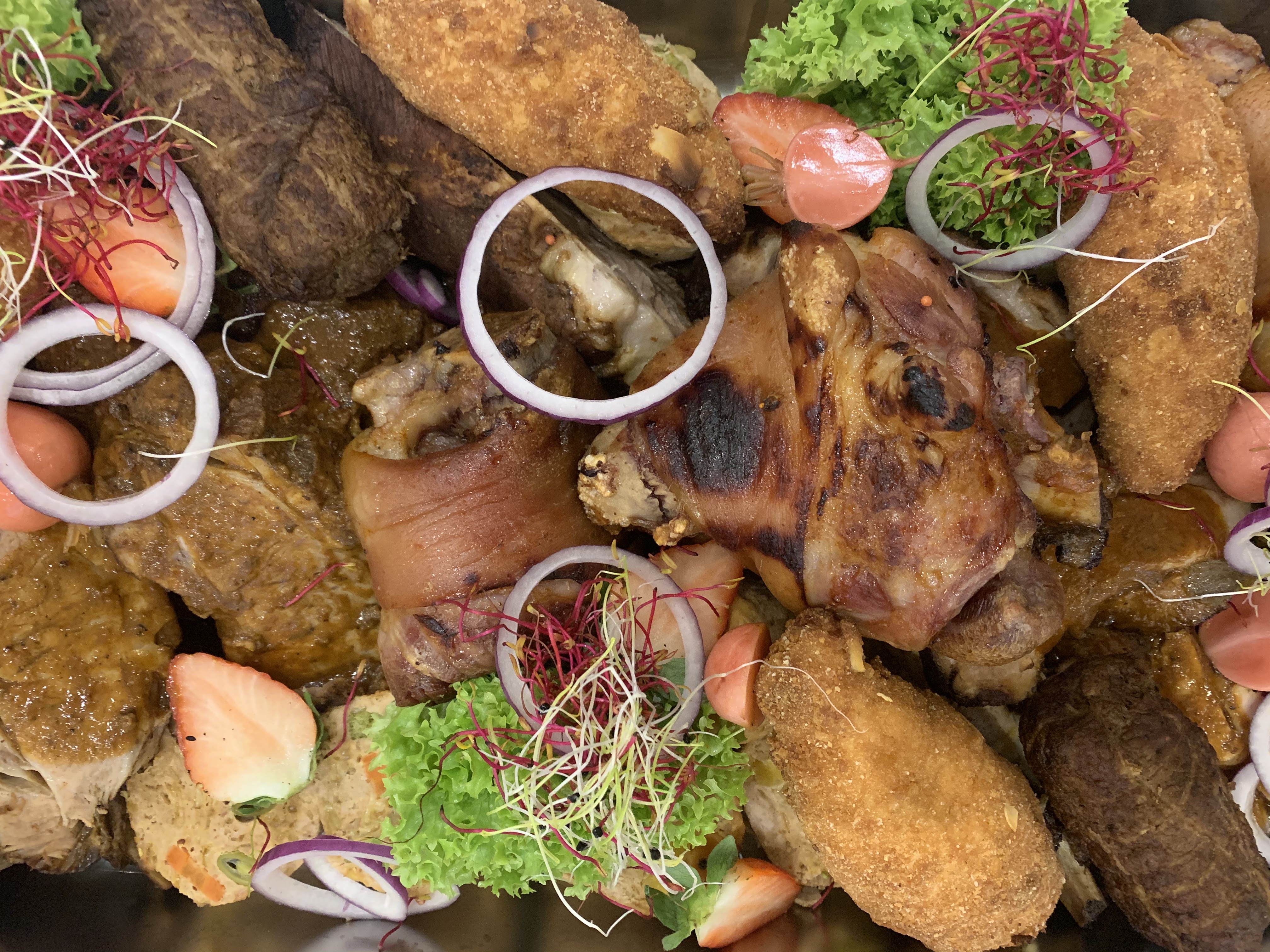 Ułożone na blasze pieczone golonki, kotlety panierowane, sałata, rzodkiewki i krążki cebuli czerwonej, udekorowane kiełkami i kawałkami truskawek.