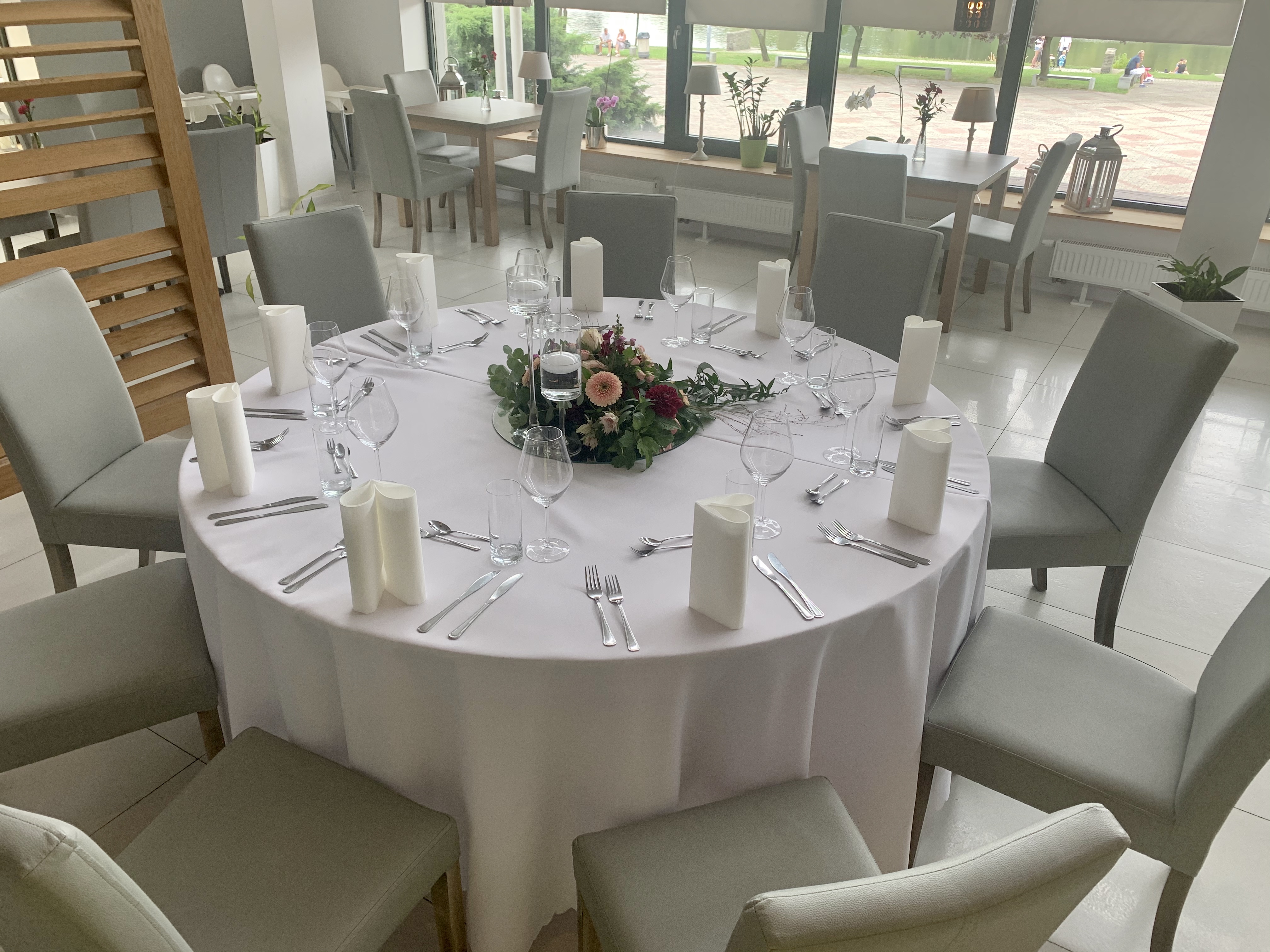 Elegancko nakryty okrągły stół z białym obrusem, szklanymi naczyniami, srebrnymi sztućcami i centralną kompozycją kwiatową, otoczony szarymi krzesłami w jasnym wnętrzu restauracji.