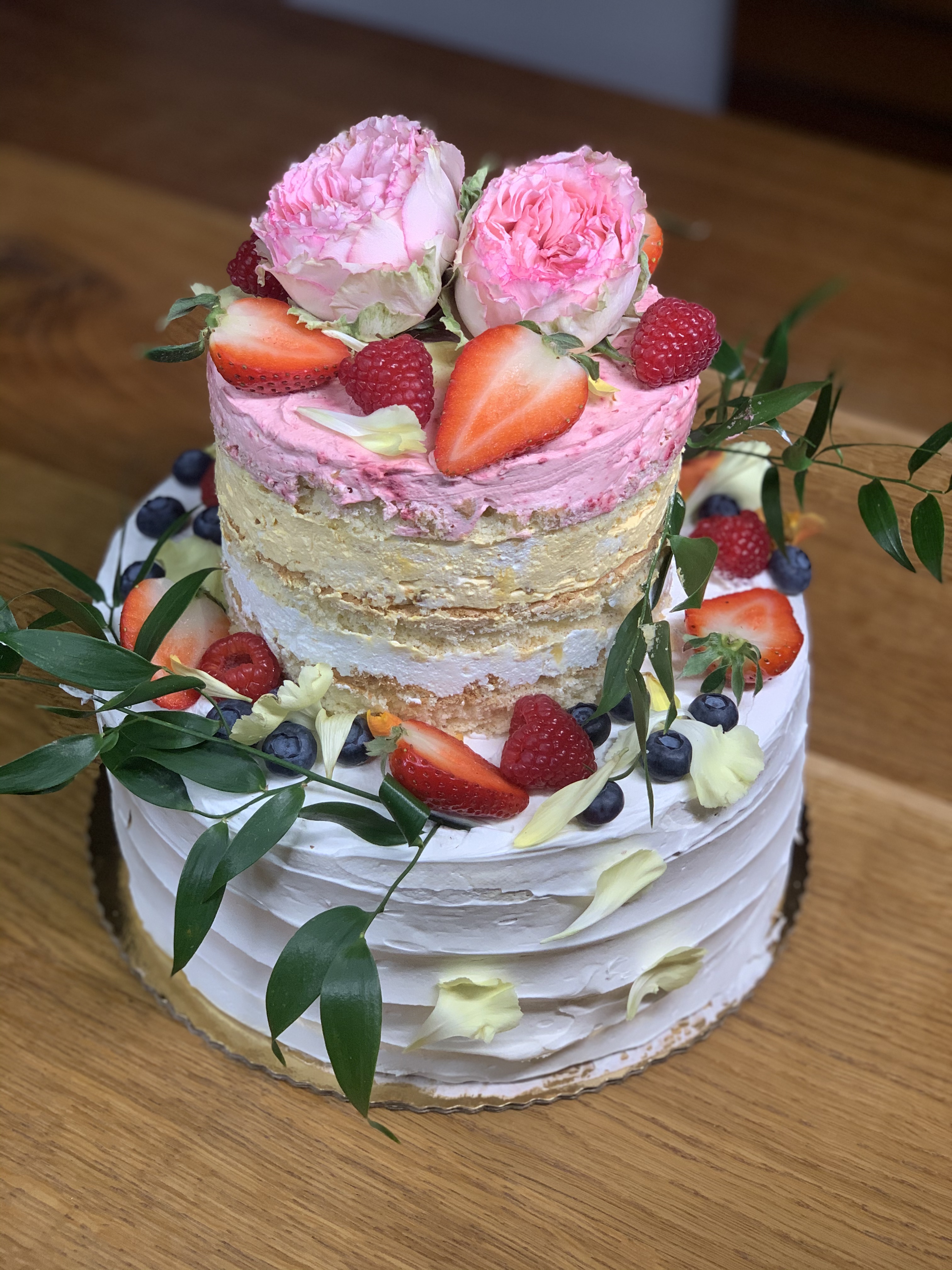 Dwupoziomowy tort z białym kremem, udekorowany truskawkami, malinami, borówkami, zielonymi listkami i dwoma różowymi różami na górze, na drewnianym blacie.
