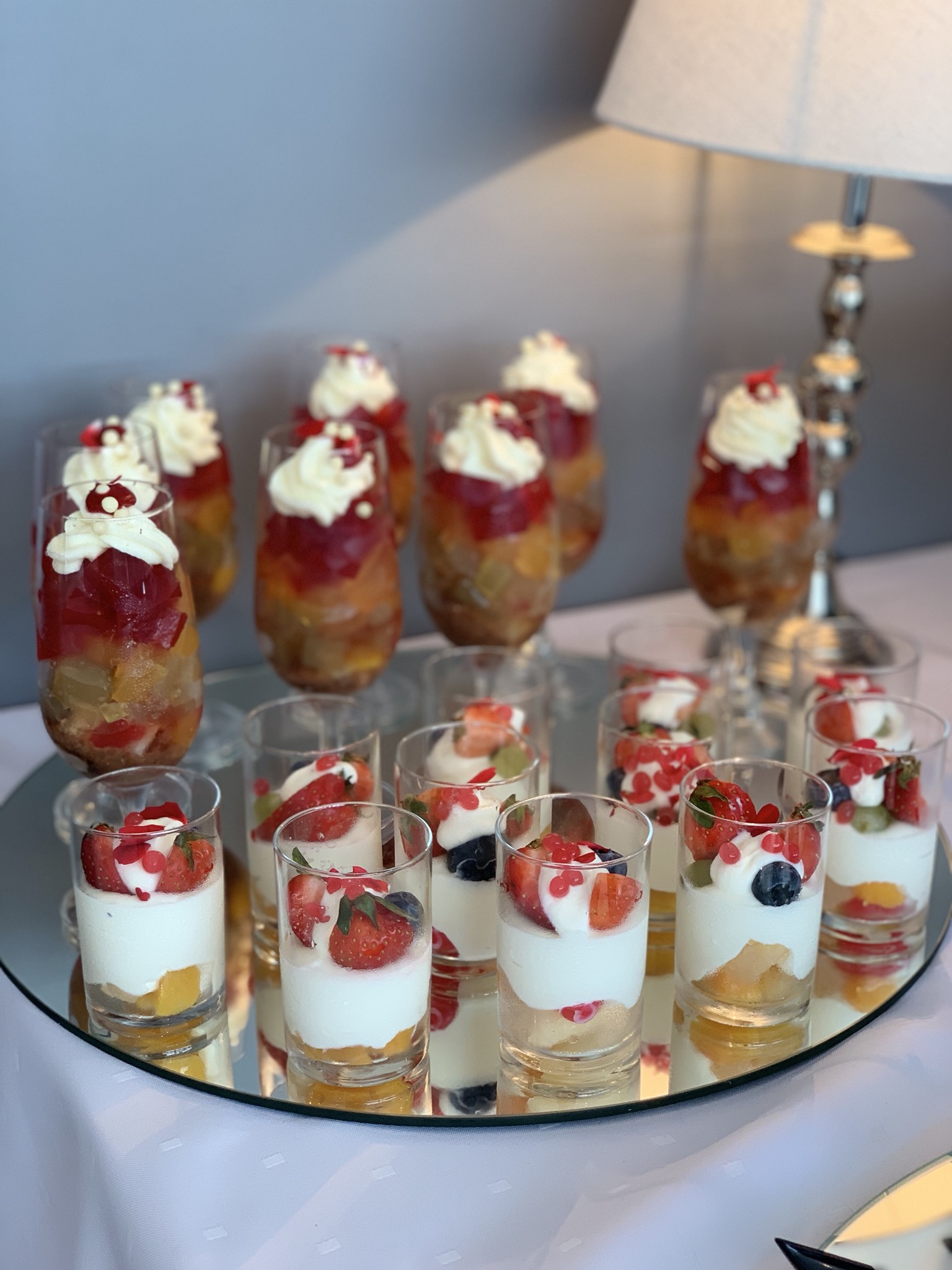 Deserki w szklankach: galaretka z owocami i bitą śmietaną oraz jogurt z owocami, przygotowane w Świętochłowicach i ustawione na okrągłym lustrze, w tle lampa stołowa.