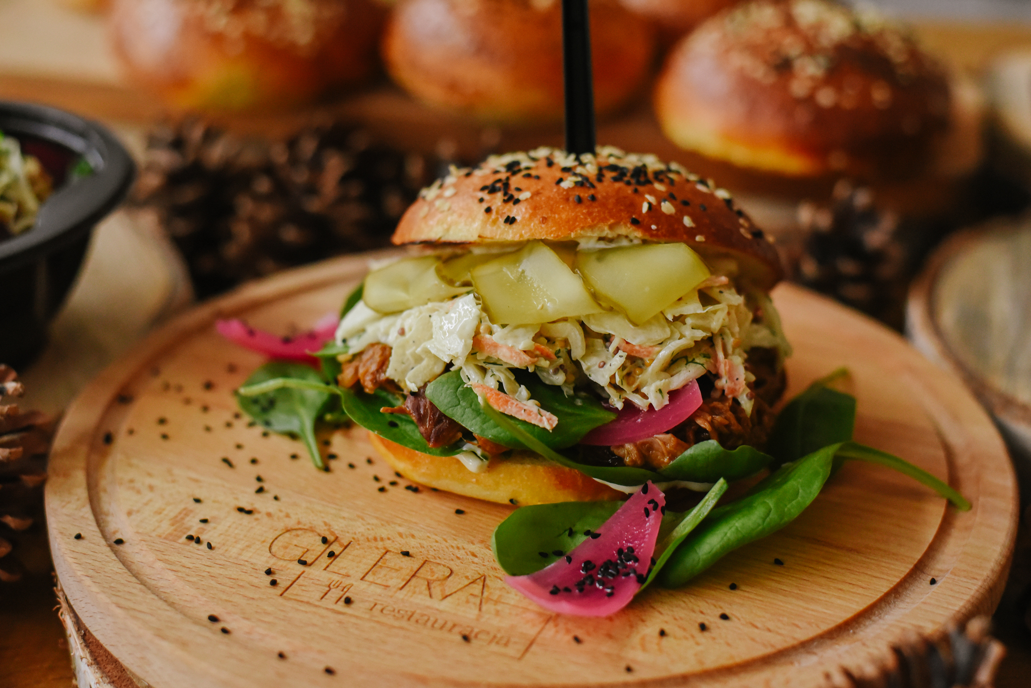 Apetyczny burger z szarpaną wieprzowiną, colesławem, piklami i marynowaną cebulą, podany na drewnianej desce z logo restauracji, w otoczeniu szyszek.