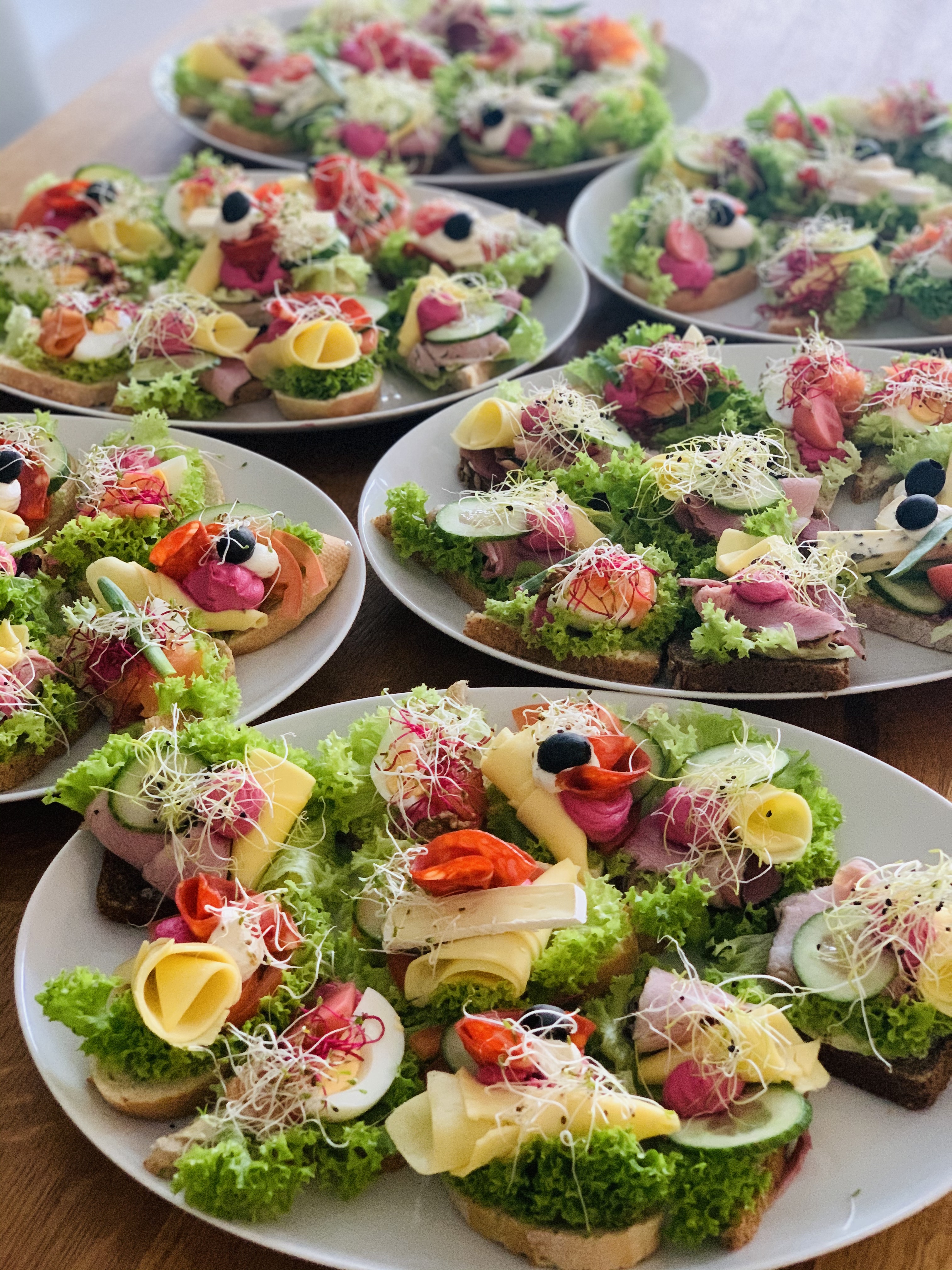 Dekoracyjne kanapki na jasnym pieczywie, ułożone na białych talerzach, z różnorodnymi dodatkami: wędlinami, serami, warzywami i kiełkami, prezentowane na drewnianym stole.