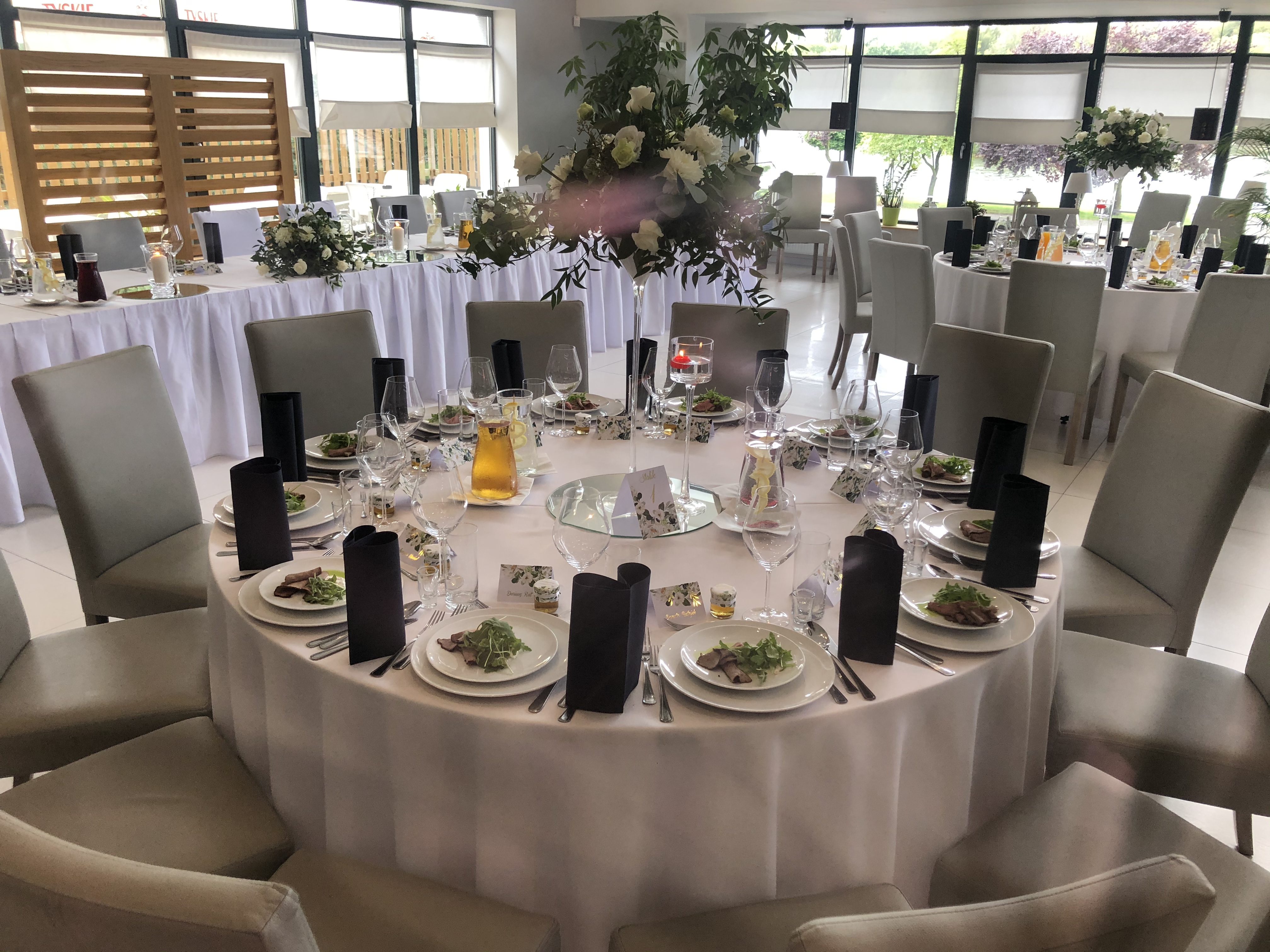 Elegancko nakryty okrągły stół z białym obrusem, szare krzesła, dania na talerzach, szklanki, czarne serwetki, w tle dekoracje kwiatowe i okna.
